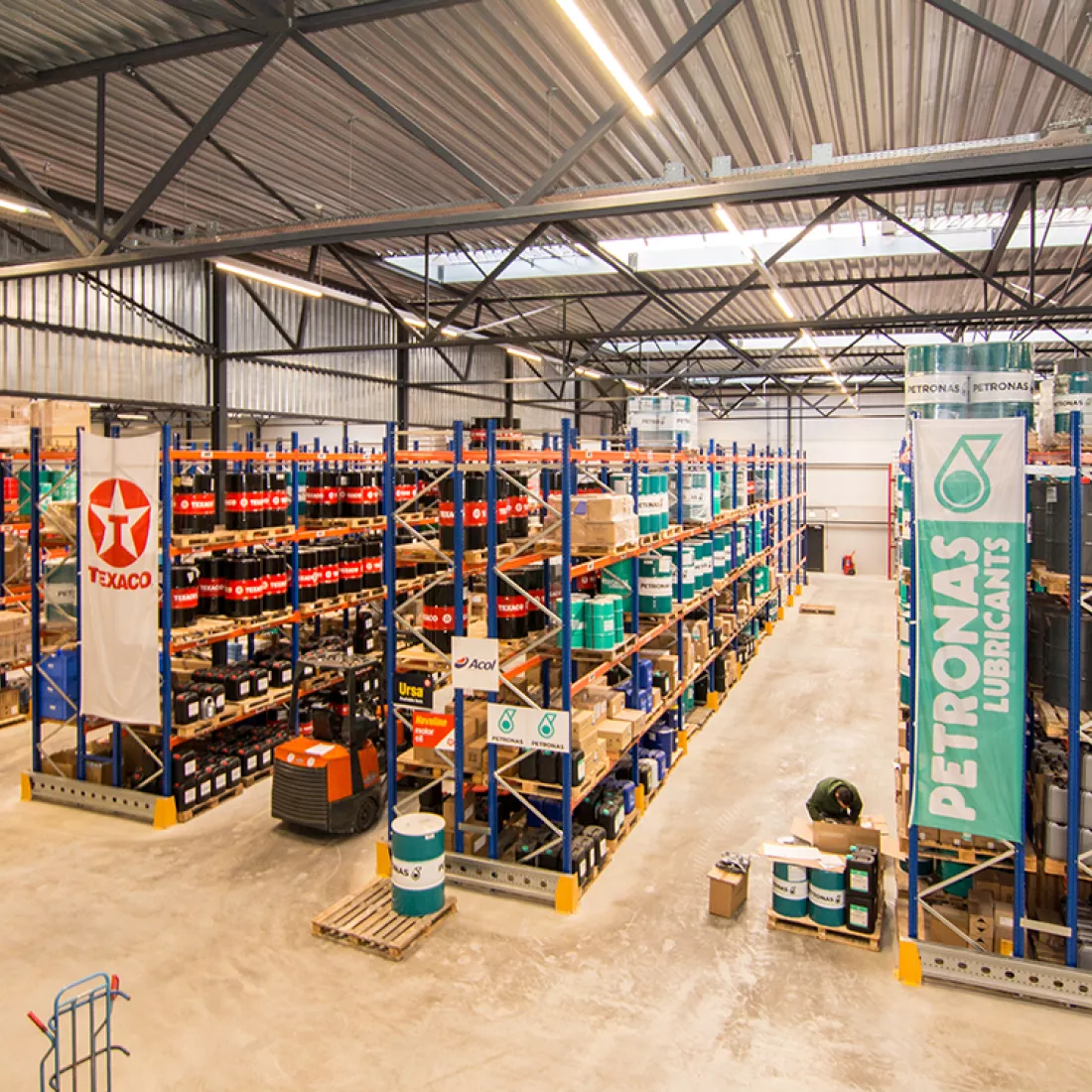 Binnenkant magazijn Acol. Er staan rijen van oranje met blauwe rekken met de producten. Het plafond bevat ook lichtstraten. Bij de opbouw is er staal gebruikt. 