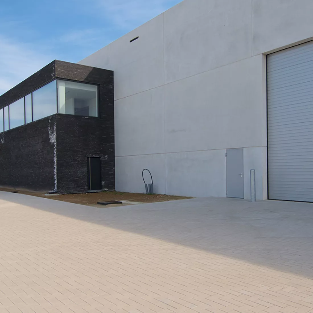 Zijaanzicht productiehal en kantoren Alcom. De productiehal is grijs en heeft ook een grijze poort. Het kantoor bestaat uit zwarte steen met grote ramen.