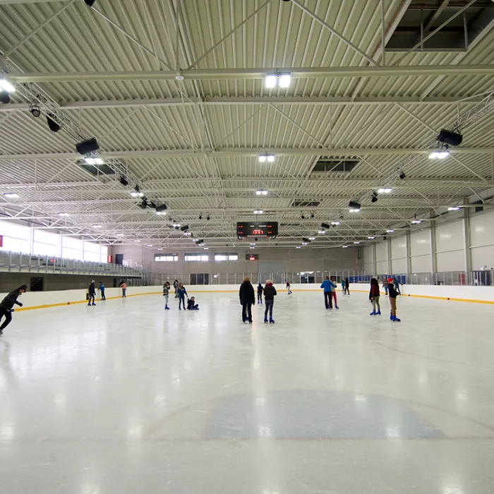 Vooraanzicht schaatspiste Ice Skating center. Er zijn mensen aan het schaatsen op het ijs. Links bevinden zich tribunes. Tegen het plafond hangen lichten.