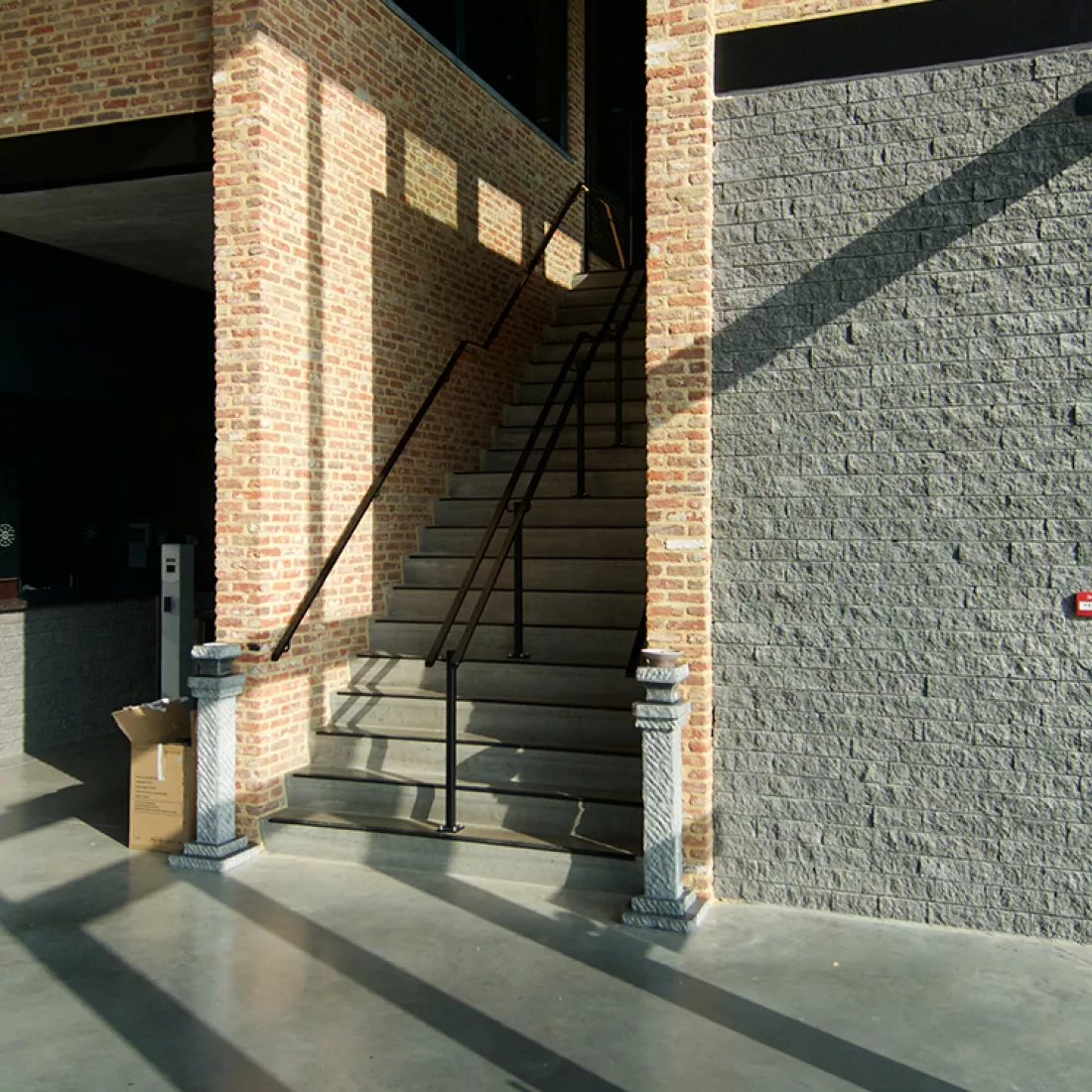 Inkom cafetaria Ice Skating center. De foto is gemaakt vanaf de onderkant van de trap. De muur rechts bestaat uit grijze steen, de andere muren uit rode steen. De trap heeft een zwarte trapleuning.