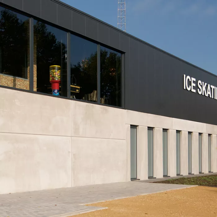 Buitenkant Ice Skating center. De onderkant van het gebouw bestaat uit beton en heeft lange smalle ramen. De bovenkant van het gebouw is zwart en heeft grotere ramen. Bovenaan hangen witte letters die het woord 'Ice Skating' vormen.