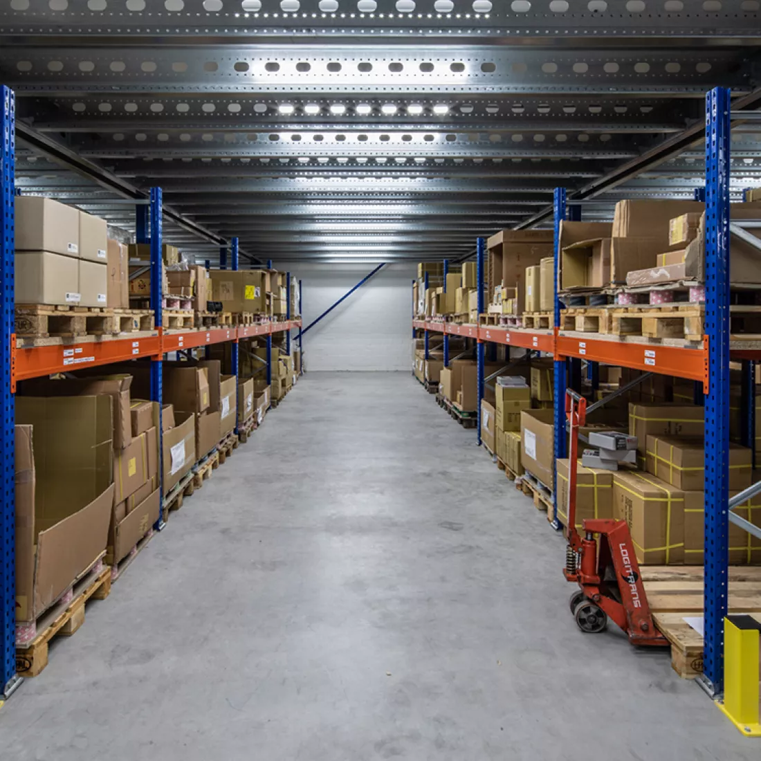 Binnenkant nieuwbouw magazijn Nauta Holding. De afbeelding is van een rij met aan beide kanten oranje met blauwe rekken. In de rekken staan producten opgeslagen in kartonnen dozen. Deze dozen staan op paletten. Onderaan rechts bevindt zich ook een transpalet.