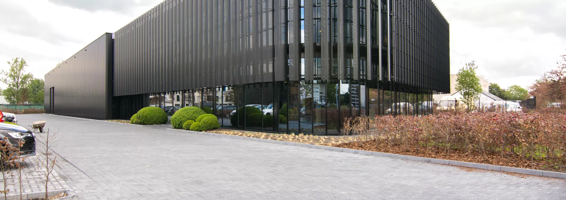 Zijaanzicht KMO gebouw met loods en kantoren van Power Solutions. het gebouw is zwart en bevat veel ramen. Rechts staat een haag. Links is een parking voor auto's. Het gebouw is gemaakt uit staal.