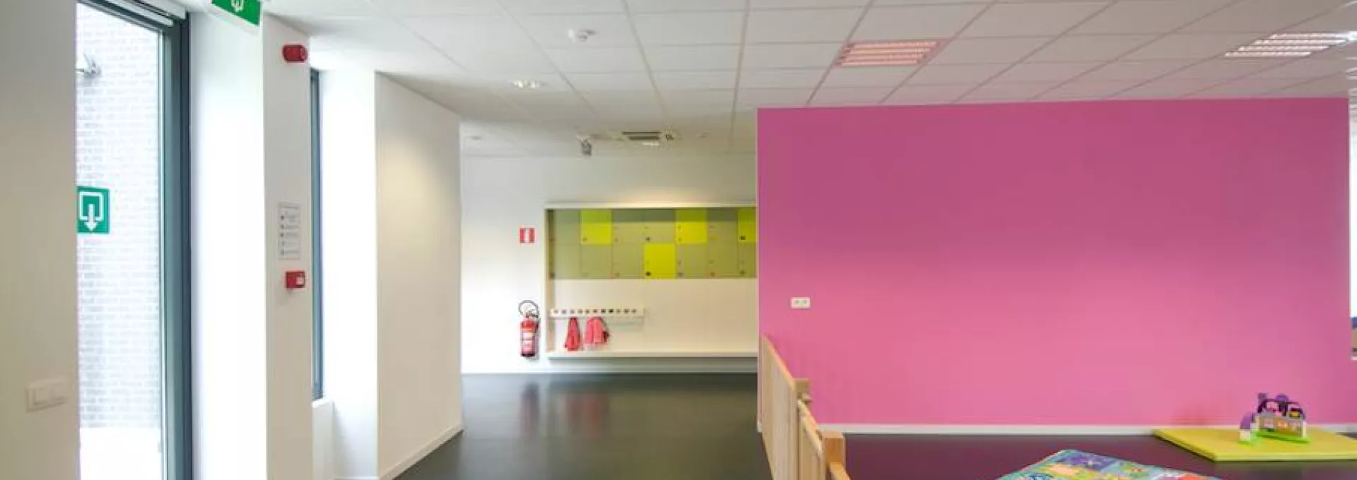 Binnenkant kinderdagverblijf. Het gebouw is opgebouwd met staal. Er is een roze muur en witte muren. Links bevinden zich twee ramen.