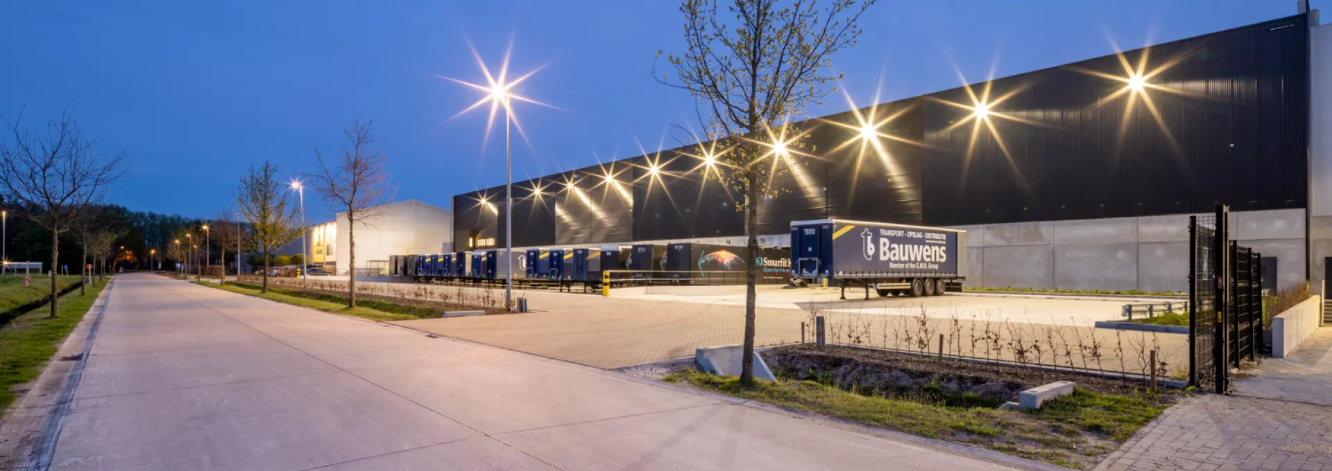 Zijaanzicht opslagmagazijn en kantoor Bauwens Logistics. Rechts bevindt zich het gebouw. Het gebouw is zwart met grijs. Links bevindt zich een straat. Voor het gebouw staan containers voor vrachtwagens. Het gebouw is opgebouwd met staal.