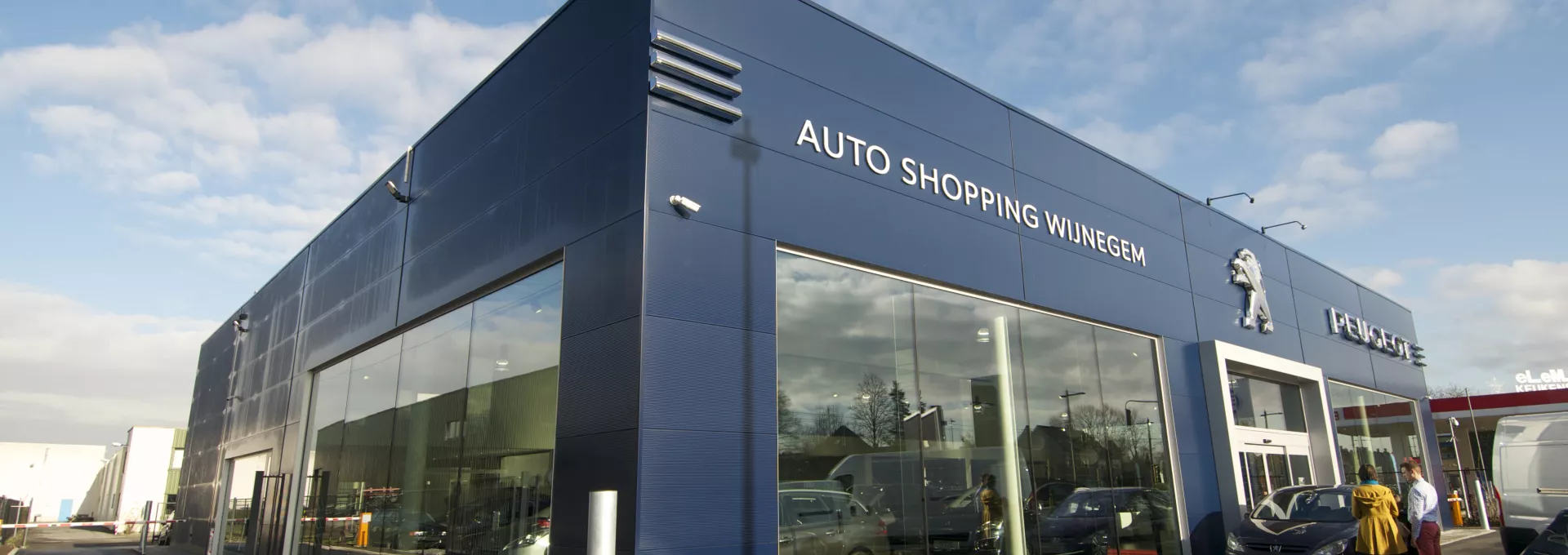 Zijaanzicht Peugeot Garage - Gabriel Car. Het gebouw is donkerblauw. Aan de voorkant hangen links bovenaan witte letters die de woorden 'shopping Wijnegem' vormen. In het midden hangt het Peugeot logo en rechts Peugeot voluit geschreven. Het gebouw heeft grote ramen om de auto's te kunnen zien. Bij de opbouw is er gebruik gemaakt van staal.