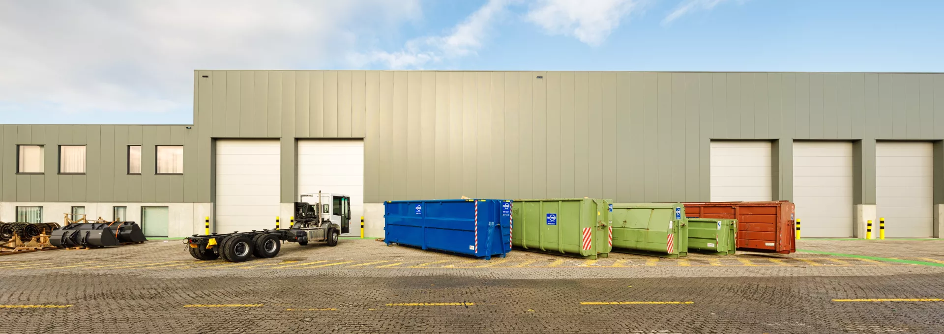 Vooraanzicht Wallenius werkplaats met bijhorende sociale ruimtes en burelen. De werkplaats bevindt zich rechts en de burelen links. Het gebouw is gemaakt met staal en heeft een grijze kleur. De burelen hebben ramen met zwarte omkadering en de de werkplaats heeft witte lange poorten. Voor de werkplaats staan verschillende containers en een truck.