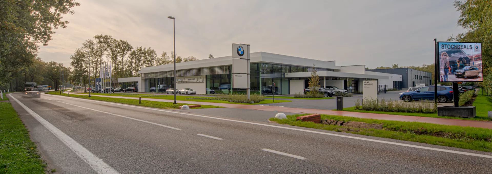 Zijaanzicht showroom garage Meeusen. De afbeelding is gemaakt vanaf de overkant van de straat. Rechts naast het gebouw zijn parkeerplaatsen voorzien. Het is een wit gebouw met grote ramen. Voor het gebouw staat een paal met drie borden. Op het bovenste bord hangt het BMW logo en op het onderste staat Meeusen.