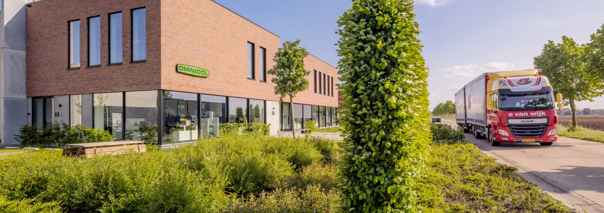 Zijaanzicht kantoor Omnicol. Links bevindt zich het kantoor en rechts een straat. Voor het kantoor staat veel groen zoals een boom. In het groen staat ook een picknicktafel. Bij de opbouw van het kantoor is staal gebruikt. De buitenkant heeft een rode steen. Onderaan zijn heel veel grote ramen. Bovenaan zijn smallere ramen. De ramen hebben een zwarte omranding. Het logo van Omnicol hangt ook tegen het kantoor.