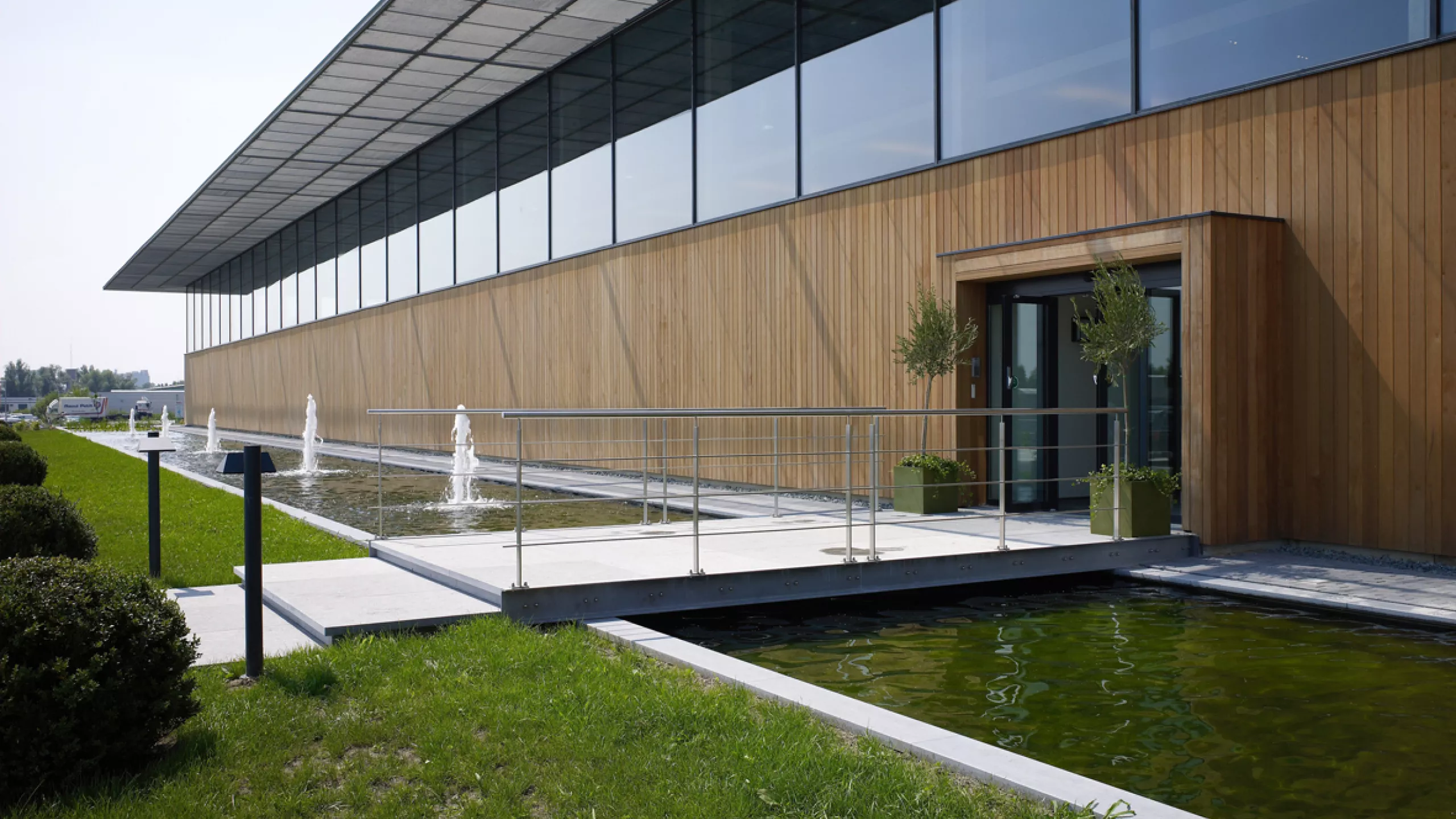 Magazijnruimte met kantoor - Ethnicraft. Het kantoor is opgebouwd uit beton en mooi afgemaakt met houten planken. Voor het gebouw is een vijver met kleine fonteinen in. Voor de inkomdeur is een kleine brug over de vijver.