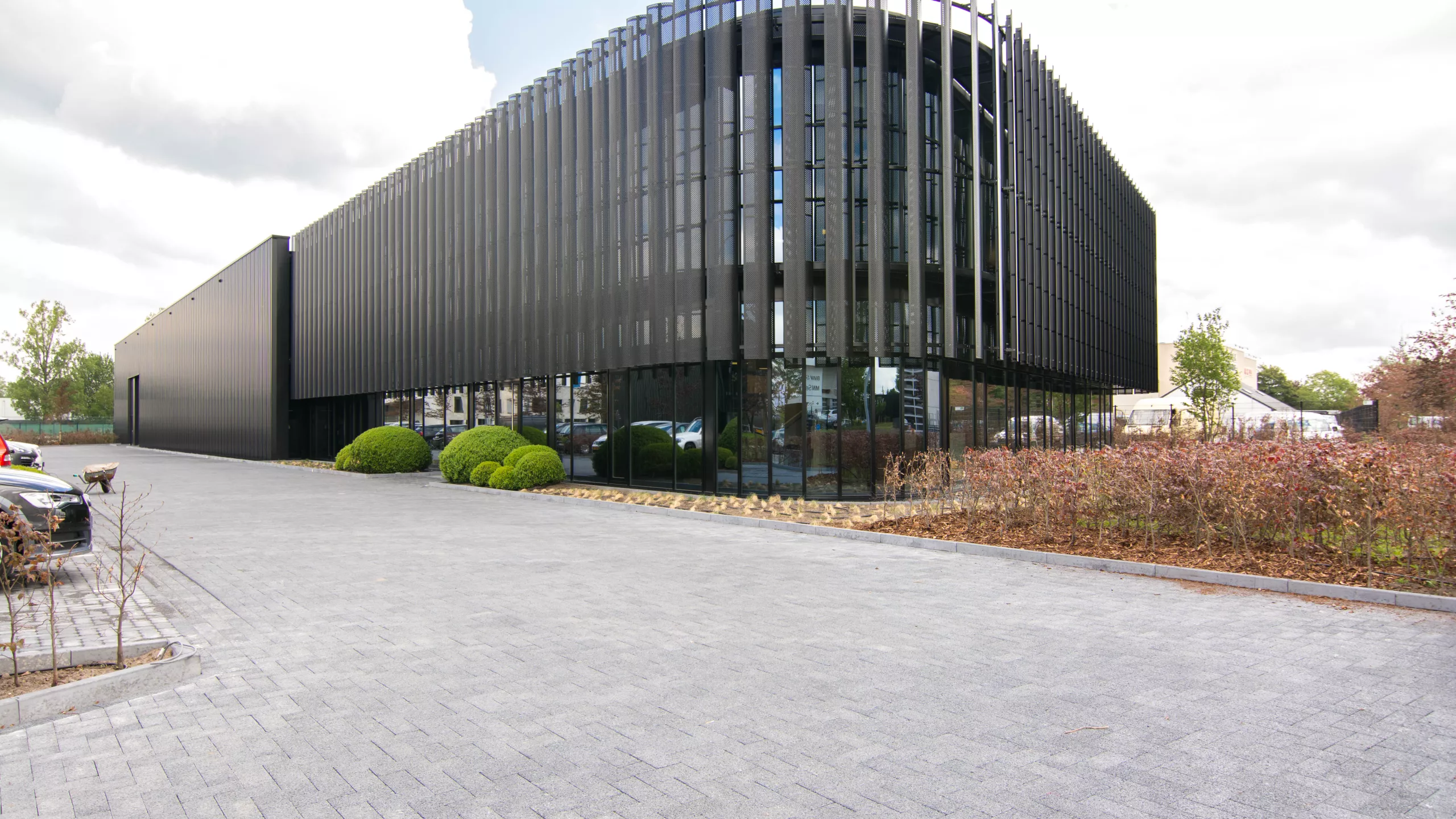 Zijaanzicht KMO gebouw met loods en kantoren van Power Solutions. het gebouw is zwart en bevat veel ramen. Rechts staat een haag. Links is een parking voor auto's. Het gebouw is gemaakt uit staal.