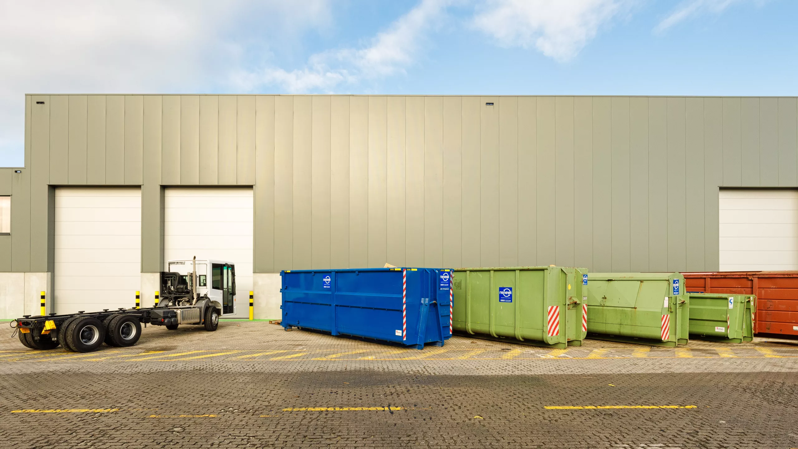 Vooraanzicht Wallenius werkplaats met bijhorende sociale ruimtes en burelen. De werkplaats bevindt zich rechts en de burelen links. Het gebouw is gemaakt met staal en heeft een grijze kleur. De burelen hebben ramen met zwarte omkadering en de de werkplaats heeft witte lange poorten. Voor de werkplaats staan verschillende containers en een truck.