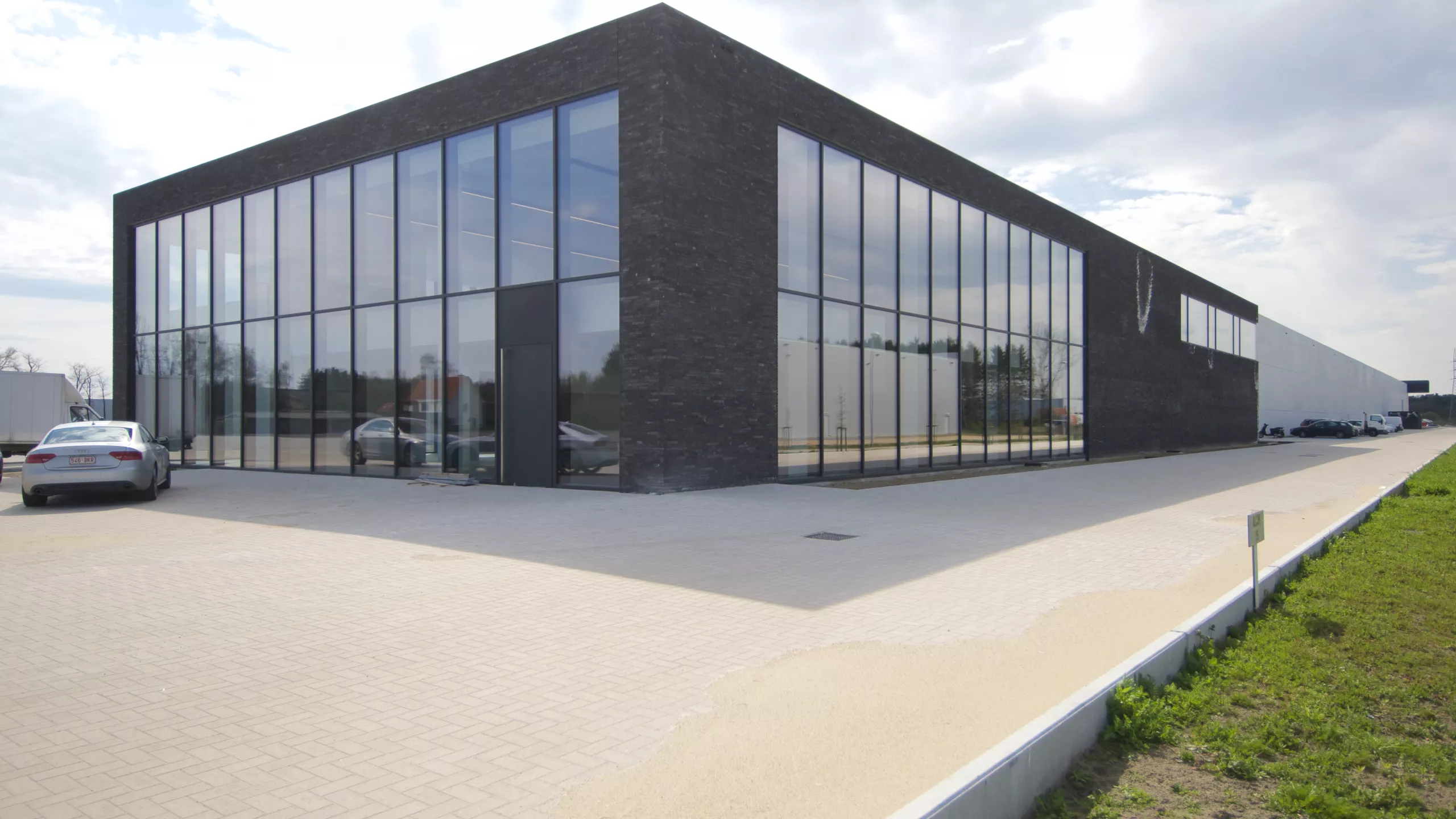 Zijaanzicht productiehal en kantoren Alcom. De productiehal is grijs en het kantoor bestaat uit zwarte steen met grote ramen. Tijdens de opbouw is er gebruik gemaakt van staal.