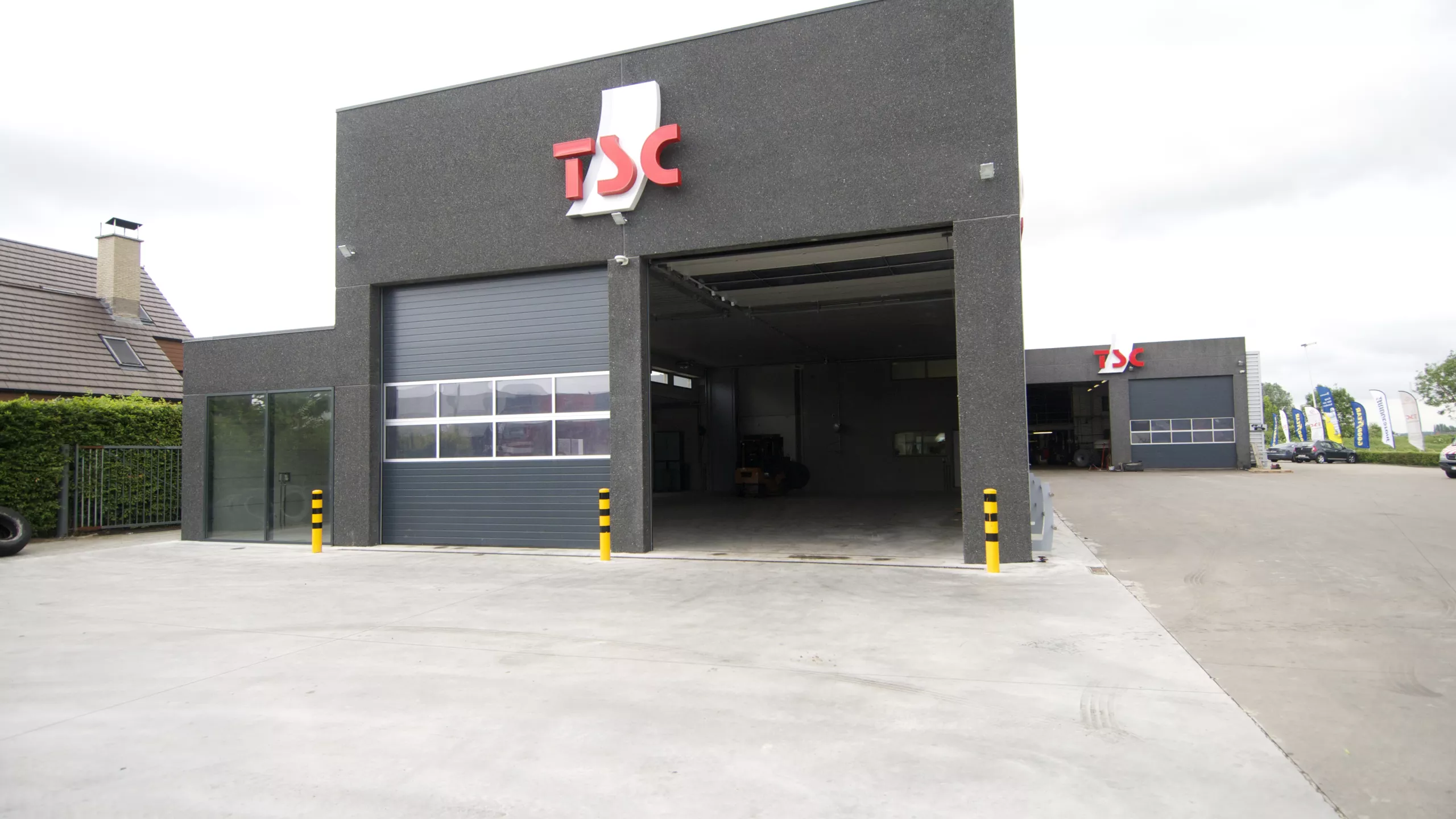 Uitbreiding opslag banden - TSC. Het gebouw is opgebouwd met een combinatie van materialen. Het gebouw is zwart langs de buitenkant en bevat ook zwarte poorten. Het logo van TSC hangt bovenaan het gebouw.