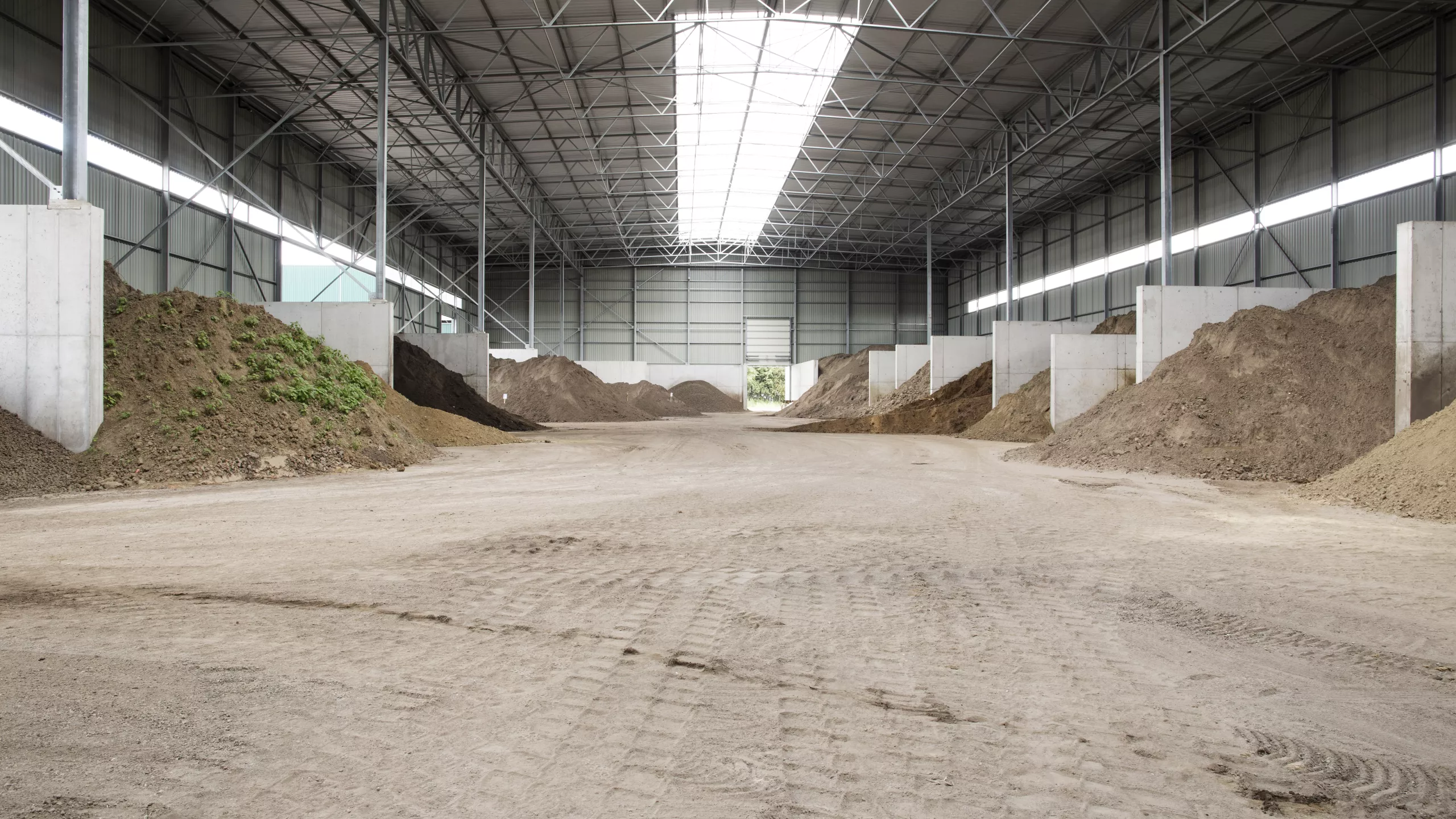 Grondhal Rumst Recycling. De hal is opgebouwd met beton. De afbeelding is langs de binnenkant genomen. Er zijn vakken verdeeld met betonnen muren.
