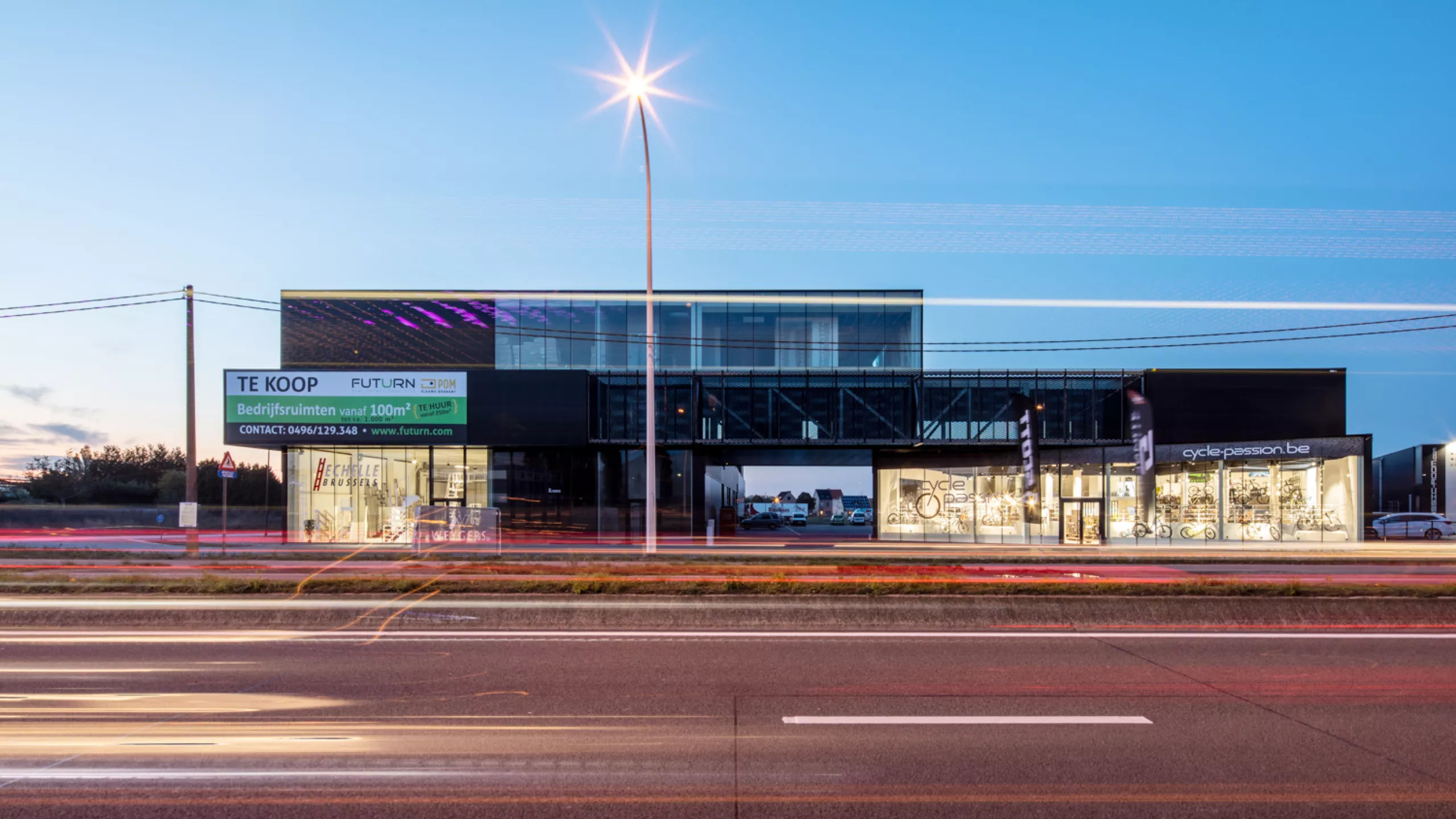 Vooraanzicht bedrijfsunits Futurn. De foto is gemaakt vanaf de overkant van de straat. Het gebouw is zwart en gemaakt uit staal. Het bevat ook veel ramen. Op de gelijkvloers is een doorgang naar een parking.