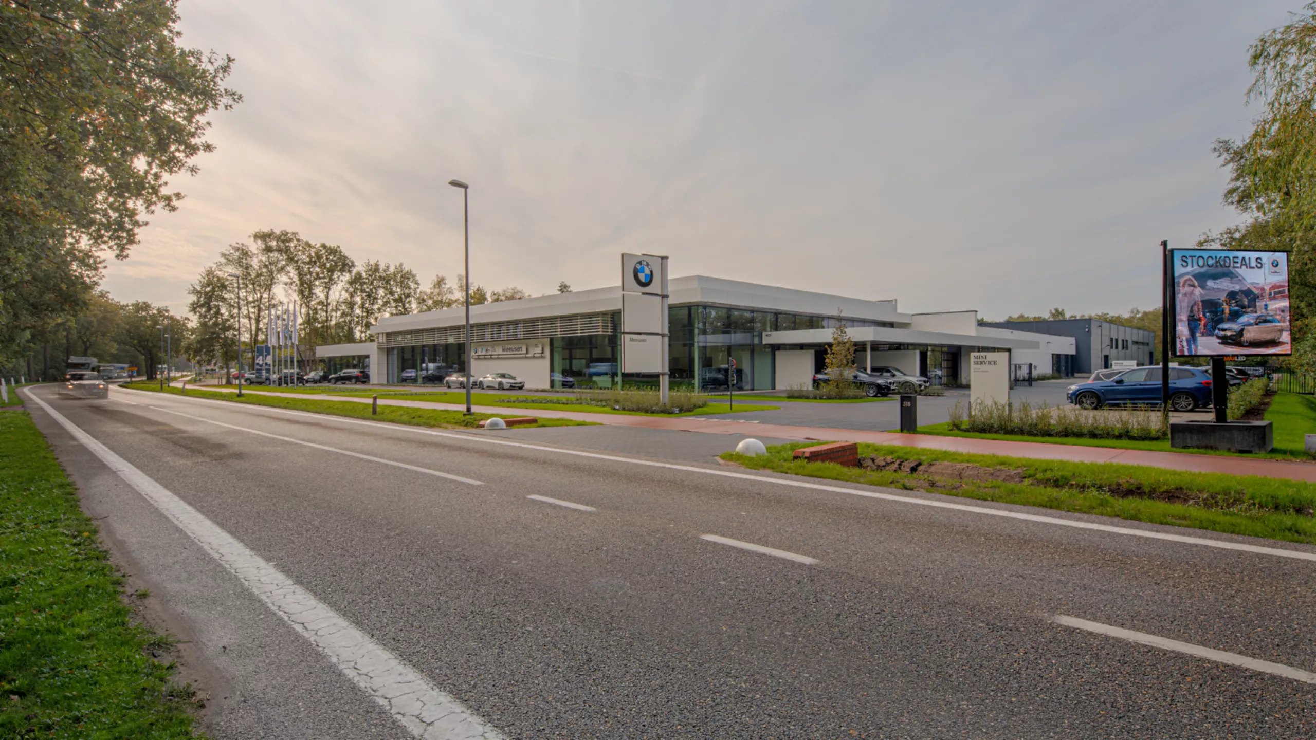 Zijaanzicht showroom garage Meeusen. De afbeelding is gemaakt vanaf de overkant van de straat. Rechts naast het gebouw zijn parkeerplaatsen voorzien. Het is een wit gebouw met grote ramen. Voor het gebouw staat een paal met drie borden. Op het bovenste bord hangt het BMW logo en op het onderste staat Meeusen.