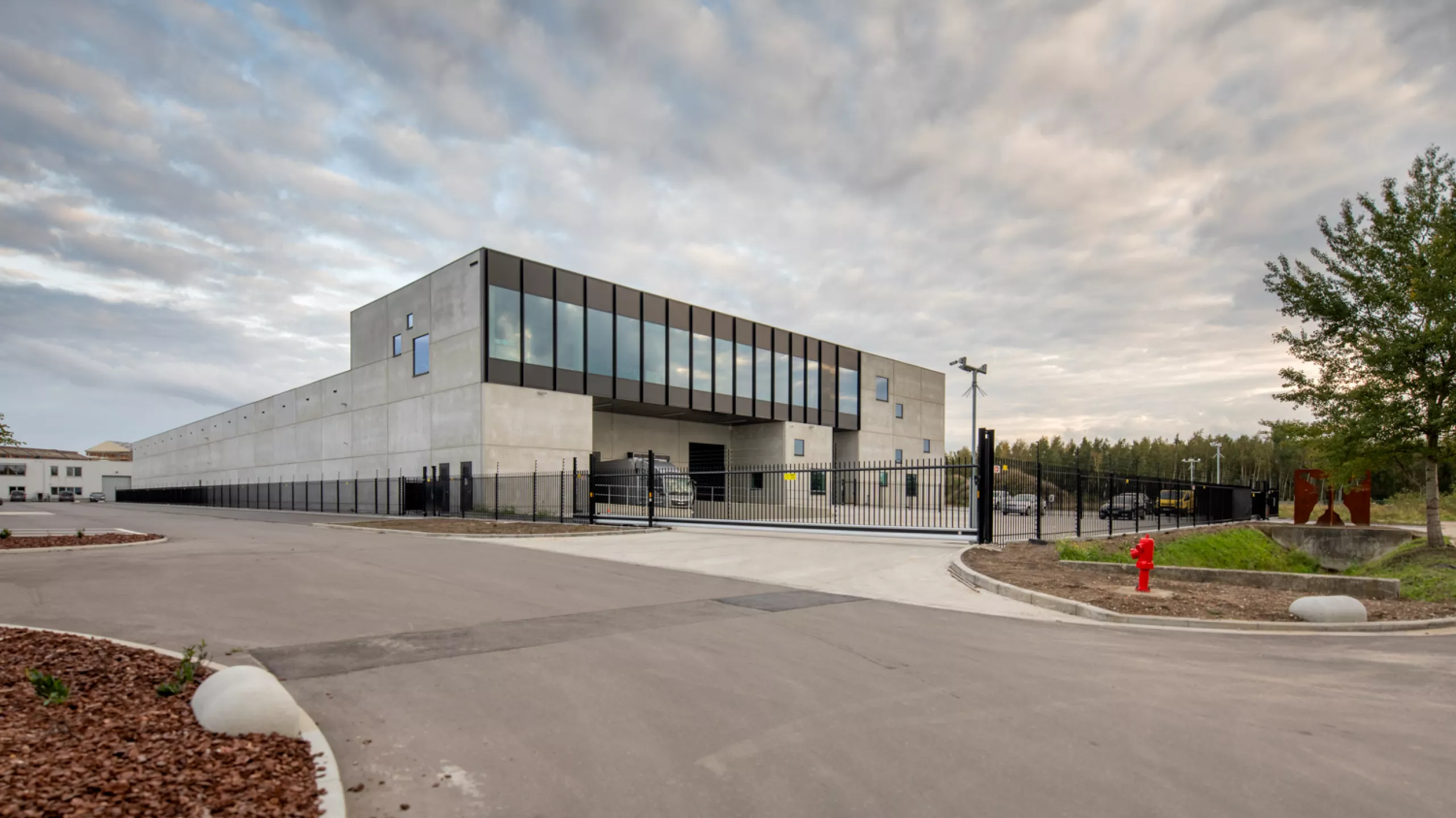 De Wille Fase 8. Bouwen van magazijn 8a + kantoor & kunstopslag. De afbeelding is een zijaanzicht van het gebouw. Het gebouw is opgebouwd met beton. Rond het gebouw bevindt zich een zwart hek met zwarte poort.