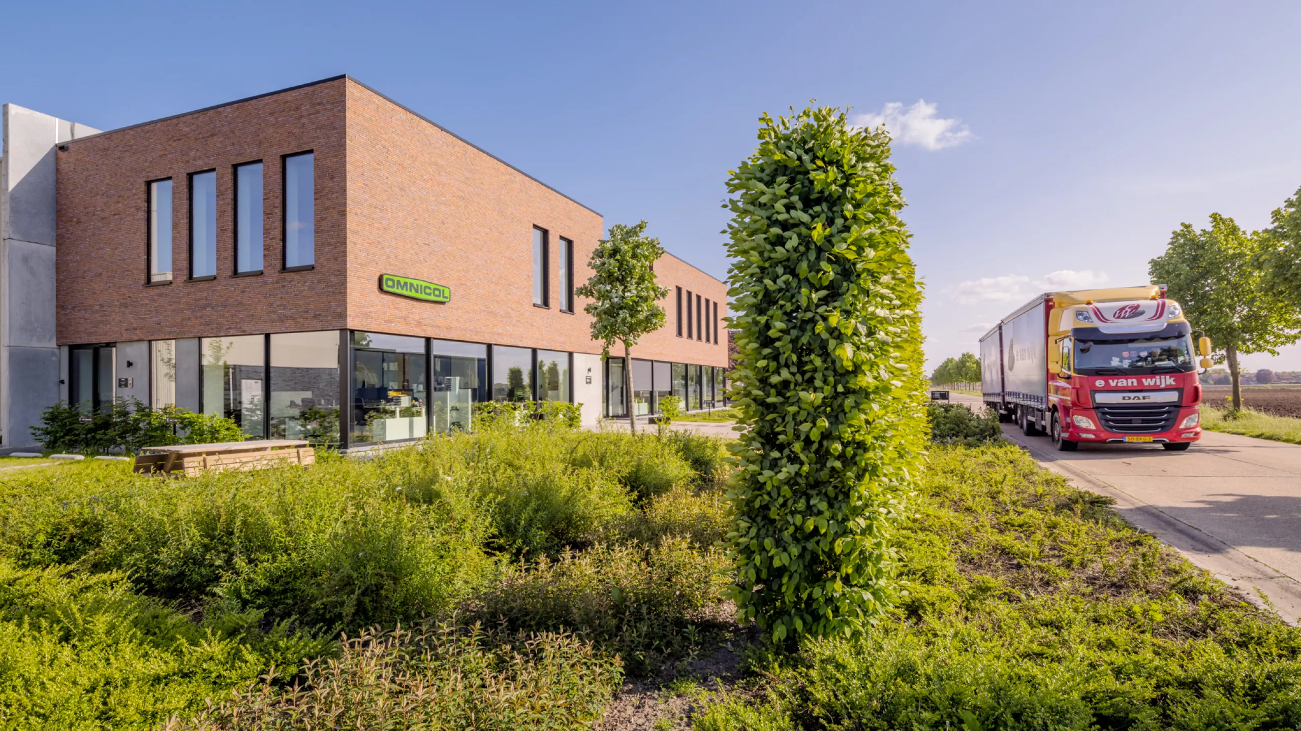 Zijaanzicht kantoor Omnicol. Links bevindt zich het kantoor en rechts een straat. Voor het kantoor staat veel groen zoals een boom. In het groen staat ook een picknicktafel. Bij de opbouw van het kantoor is staal gebruikt. De buitenkant heeft een rode steen. Onderaan zijn heel veel grote ramen. Bovenaan zijn smallere ramen. De ramen hebben een zwarte omranding. Het logo van Omnicol hangt ook tegen het kantoor.