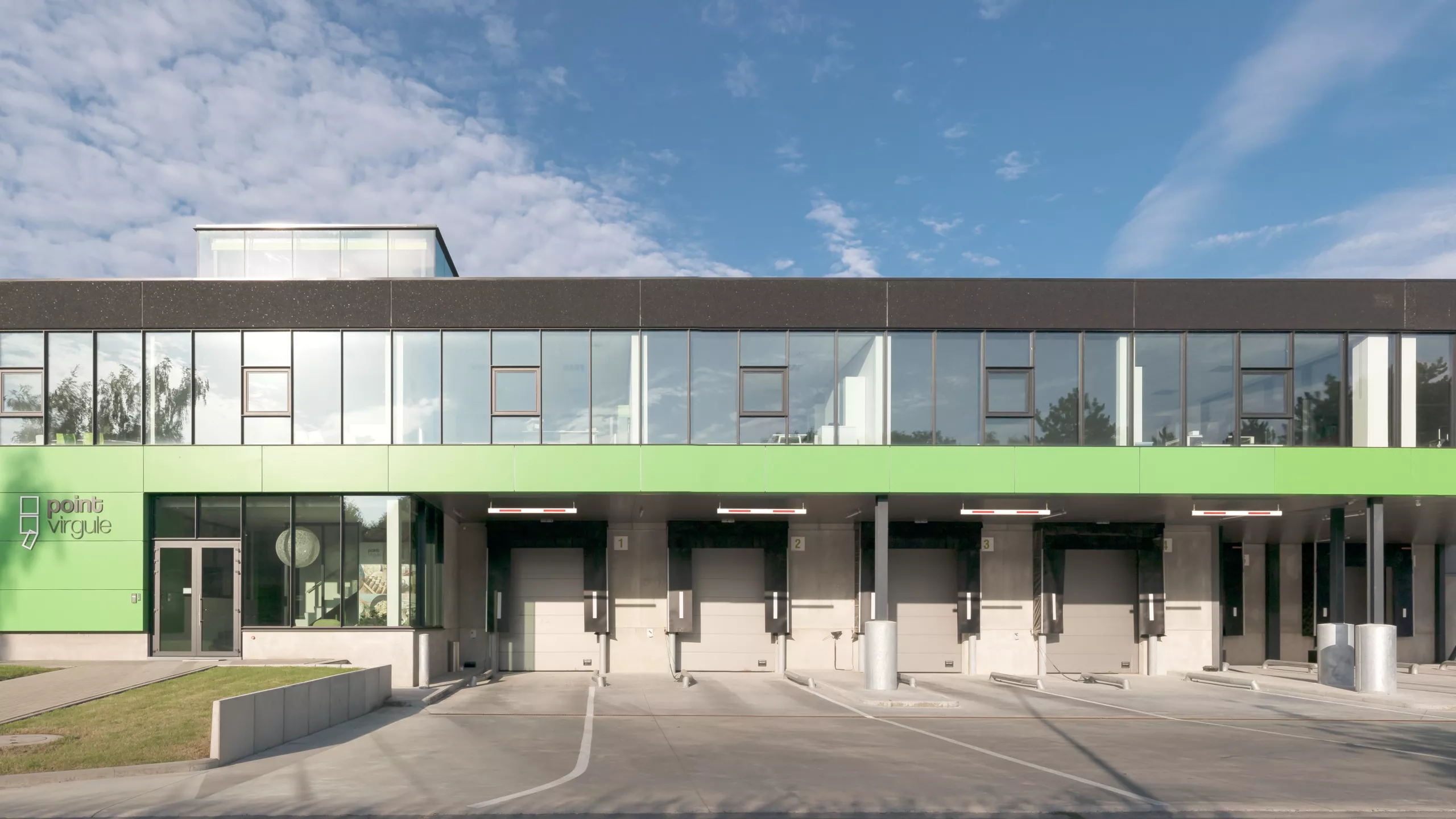 Vooraanzicht stapelmagazijn en toonzaal van Point Virgule. Het gebouw bestaat uit de kleuren grijs, zwart en groen. Het gebouw bevat zwarte stalen ramen. Rechts onderaan bevindt zich een dock voor het laden en lossen van vrachtwagens. Het gebouw is gemaakt uit staal.