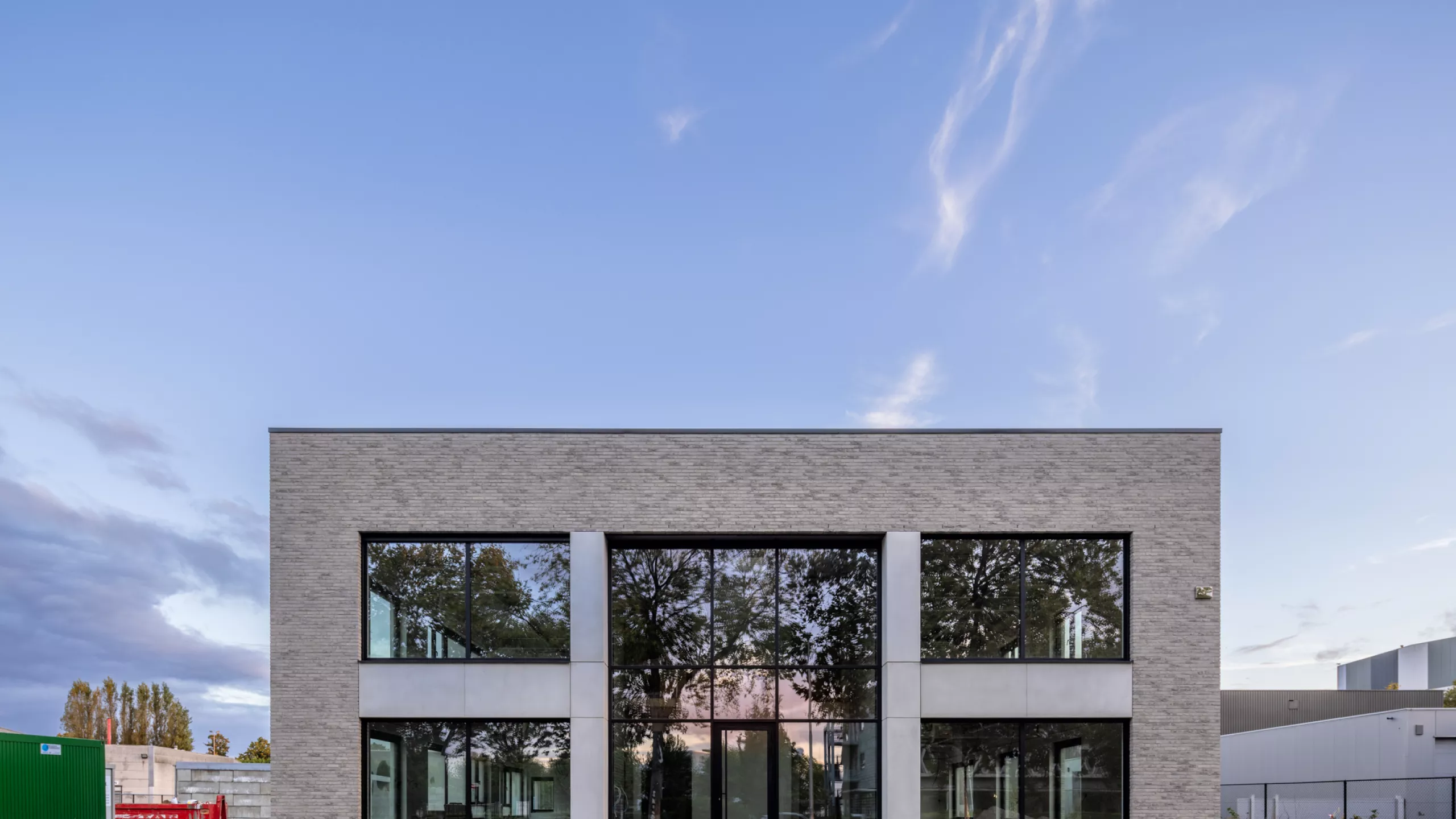 Renovatie magazijnen en kantoor L&C Trading. De afbeelding is een vooraanzicht van het kantoor. Het bestaat uit grijze stenen en beton. Ook bevat het kantoor enkele grote ramen, deze hebben een zwarte omranding.