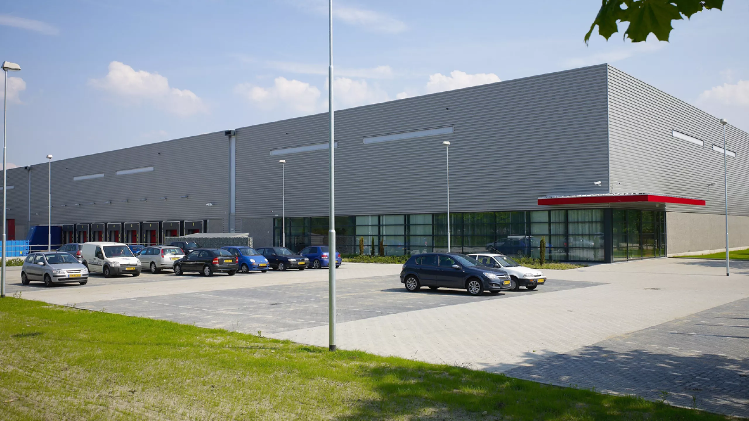 Vooraanzicht distributiecentrum van Prologis Etten Leur.  Het is een een grijs gebouw met rode details en zwarte ramen. Voor het gebouw zijn parkingplaatsen voorzien.