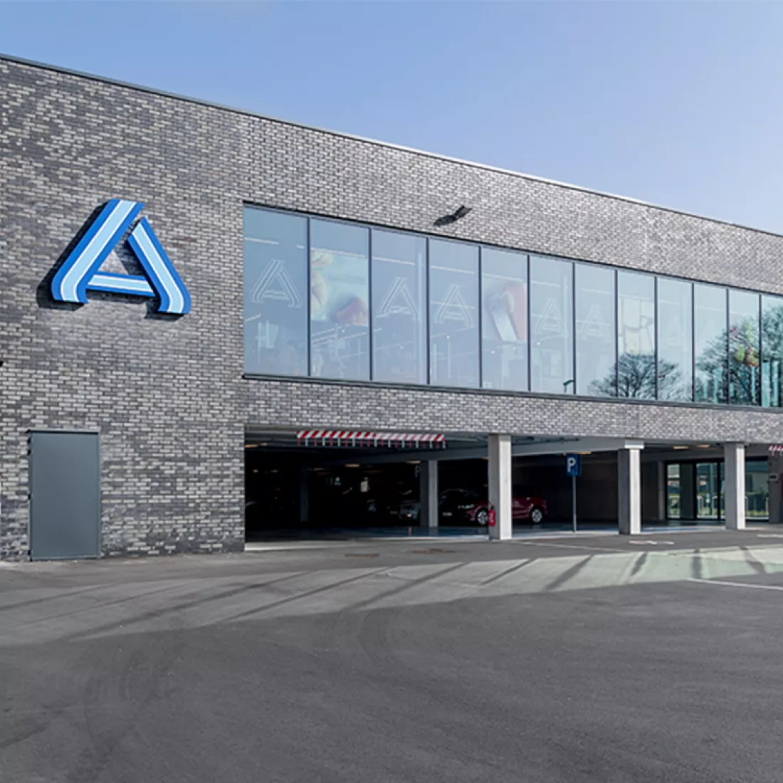 Vooraanzicht winkel Aldi Essen. Het gebouw bestaat uit grijze stenen en beton. De winkel heeft twee verdiepen. Op de gelijkvloers bevindt zich de parking. Daarboven bevindt de winkel zich. De winkel bevat veel ramen. Links naast de ramen hangt het logo van Aldi.