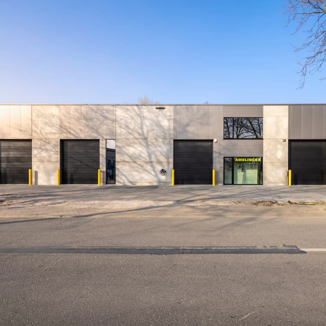 Vooraanzicht bedrijfsunits - Amelinckx Hoboken. Het gebouw bestaat uit beton en grijze platen. Het bevat zwarte poorten en verschillende soorten ramen. Voor de poorten staan gele paaltjes.