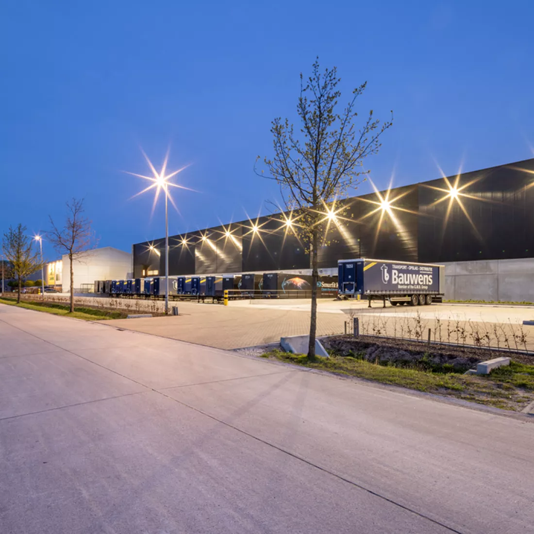 Zijaanzicht opslagmagazijn en kantoor Bauwens Logistics. Rechts bevindt zich het gebouw. Het gebouw is zwart met grijs. Links bevindt zich een straat. Voor het gebouw staan containers voor vrachtwagens. Het gebouw is opgebouwd met staal.