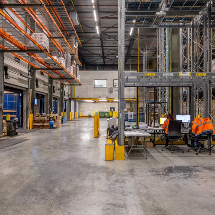 Binnenkant opslagmagazijn Bauwens Logistics. Het gebouw is opgebouwd met staal. Er staan voornamelijk rekken in het magazijn, zoals links hangen rekken tegen de muur met materiaal. Er is ook een bureau aanwezig, deze staat rechts.