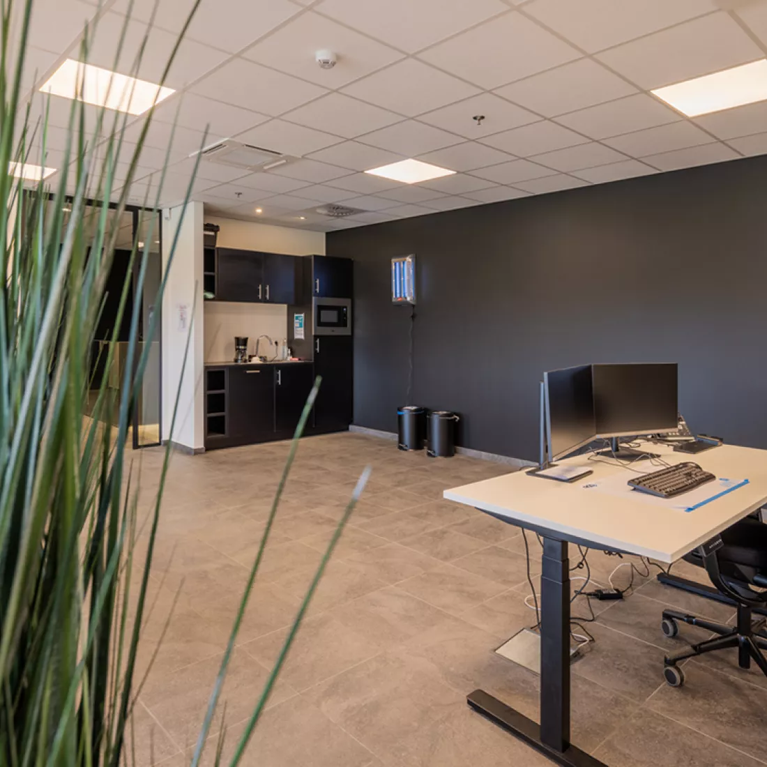 Binnenkant kantoor Bauwens Logistics. Links is er een klein keukenblok voorzien, deze heeft een zwarte kleur. De muur rechts daarvan is zwart. Rechts staat er ook een bureau.