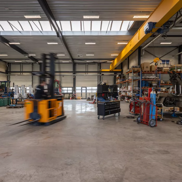 Binnenkant magazijn in het bedrijfspand van Terberg Gent. Het plafond bevat een lichtstraat. De achterste muur heeft enkele poorten. In het magazijn staan verschillende materialen en heftrucks.