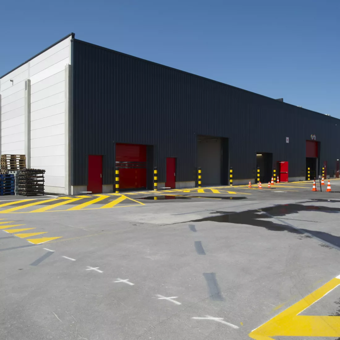 Zijaanzicht loods ICO. De zijkant van het gebouw is wit, de voorkant is zwart. Het gebouw bevat rode poorten en deuren.