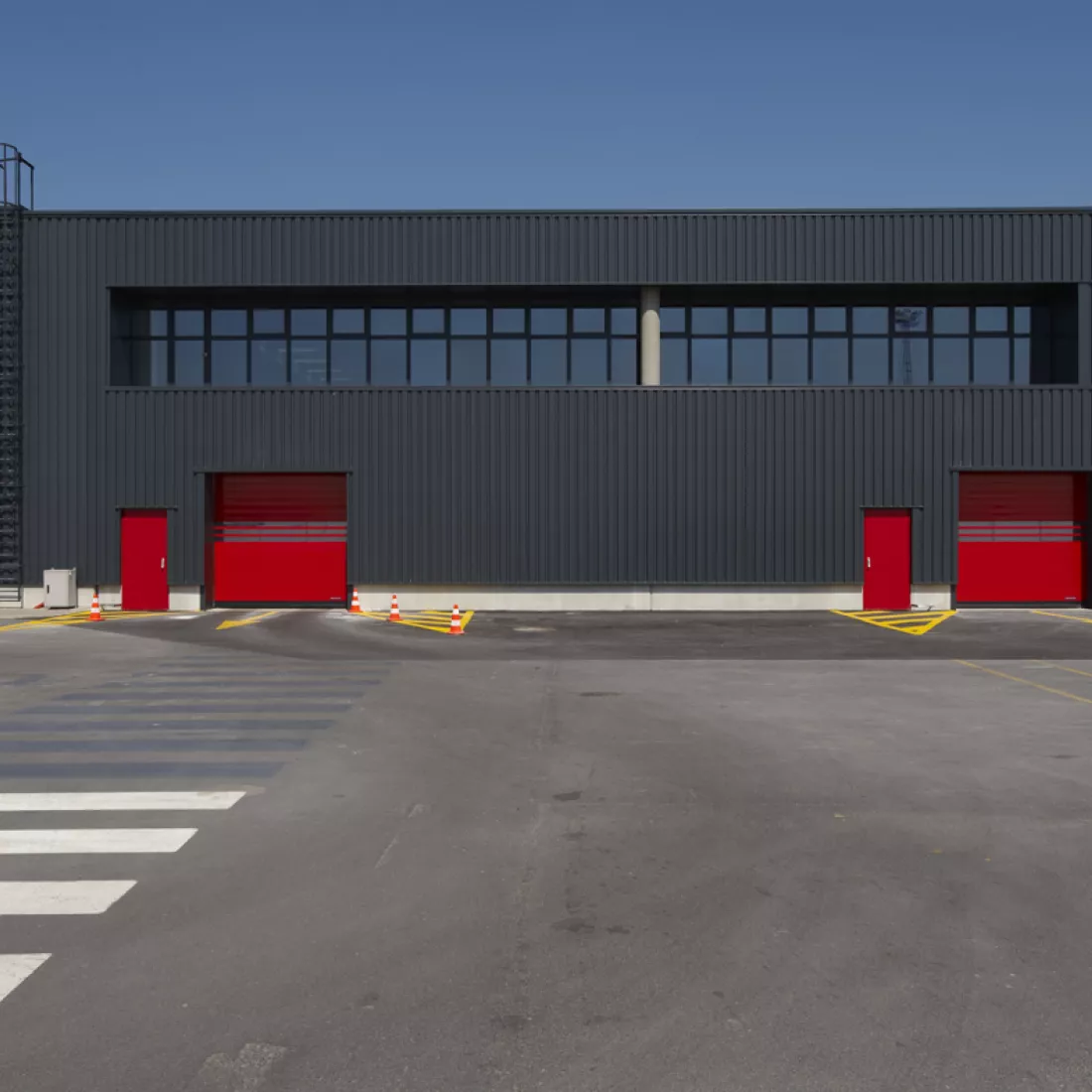Vooraanzicht loods ICO. De loods bestaat uit twee verdiepingen. De bovenste verdieping bevat veel ramen. Het gebouw is zwart, maar bevat rode poorten en deuren.