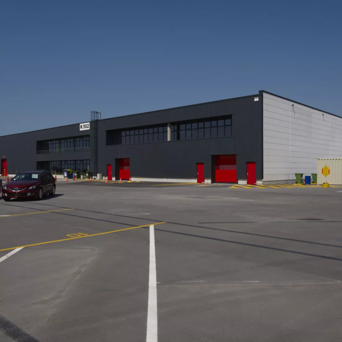 Zijaanzicht loods ICO. De zijkant van het gebouw is wit, de voorkant is zwart. Het gebouw bevat rode poorten en deuren.