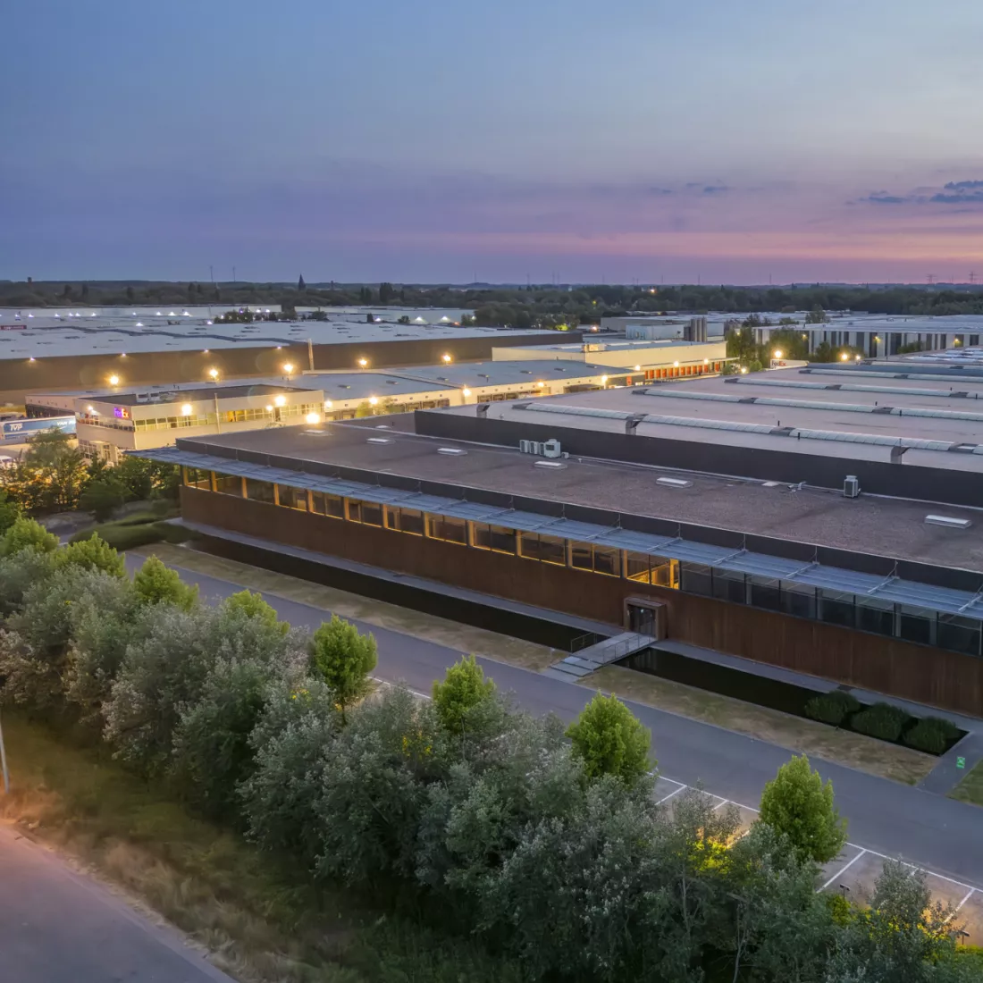 Een luchtfoto van een moderne industriële faciliteit tijdens de schemering. Het uitgestrekte gebouw met plat dak heeft talrijke dakramen en luchtunits, en is omgeven door weelderig groen en een goed onderhouden landschap. De gevel van het gebouw is deels bekleed met hout, waardoor het geïntegreerd wordt met de natuurlijke omgeving. De lucht is geschilderd met zachte tinten paars en blauw, wat de overgang van zonsondergang naar avond suggereert.
