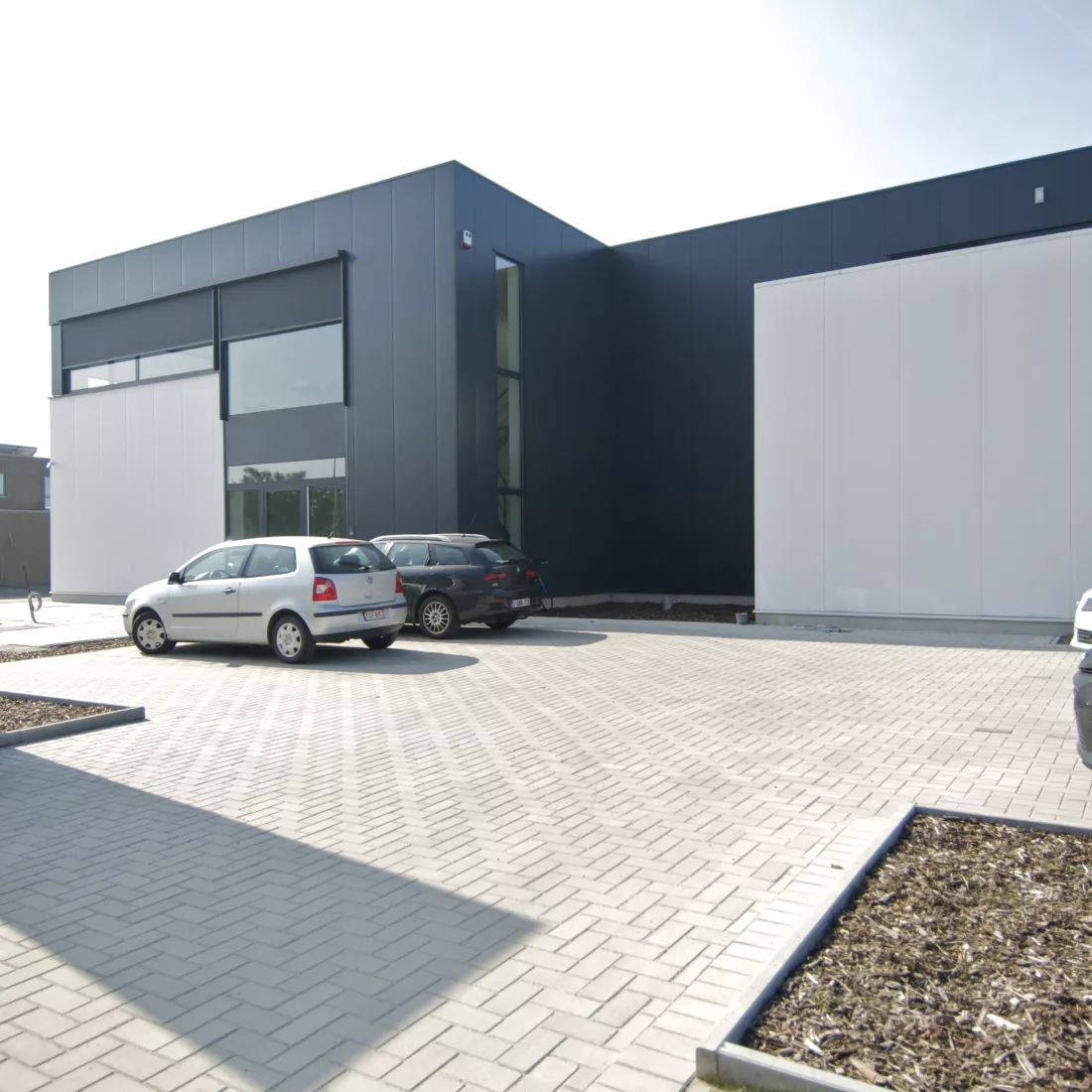 Vooraanzicht productiehal en kantoor Argina. Het gebouw is wit met zwart en bevat enkele ramen. Voor het gebouw zijn parkeerplaatsen voorzien.