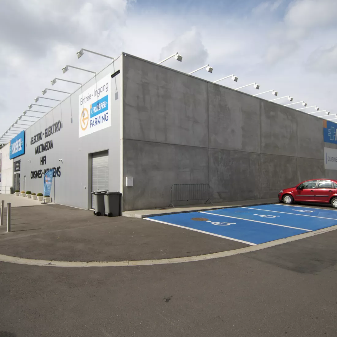 zijaanzicht van de buitenkant van de showroom. Rechts zijn parkeerplaatsen voor mensen met een beperking. Het gebouw is grijs en is gemaakt met staal.