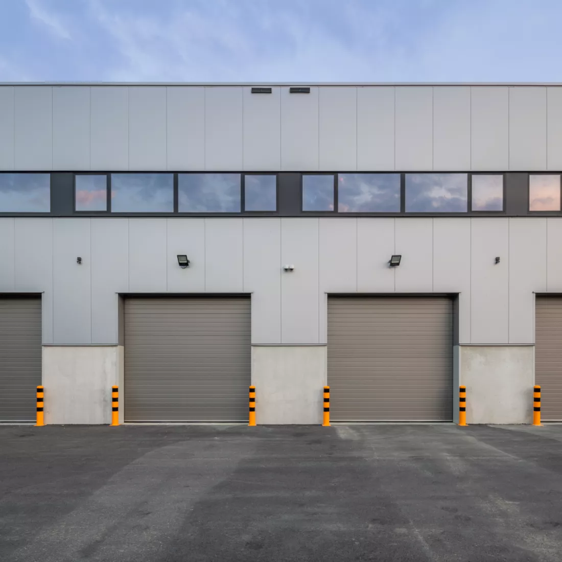 Close-up vooraanzicht magazijn Foriver. Vooraan zijn er vieer grijze poorten met daarboven ramen afgewerkt met zwart. Het gebouw is grijs en gemaakt met staal.