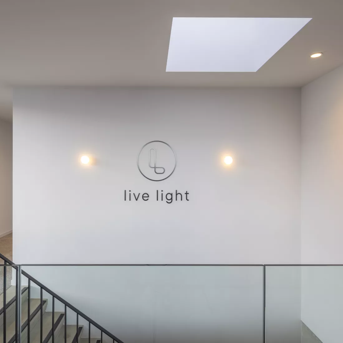 Binnenkant Ethnicraft - traphal. Achteraan bevindt zich een trap uit beton met een zwarte railing. Bovenaan is er een glazen railing rond de trap. Tegen de achterste muur hangen twee lampen en de woorden 'live light'.