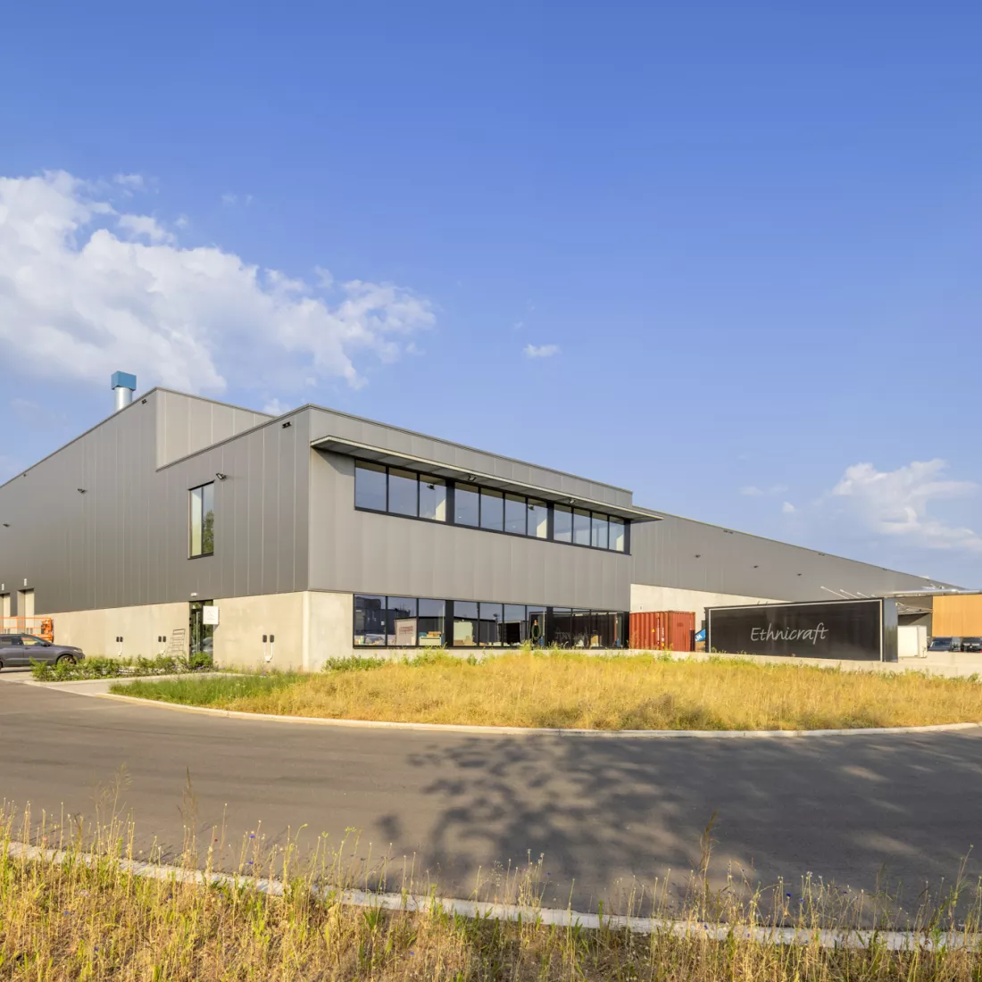 Zijaanzicht Ethnicraft. Het gebouw bestaat uit beton en staal. Aan de onderkant heeft het een betonlook en aan de bovenkant zijn grijze platen. Rechts bevindt zich een dock.
