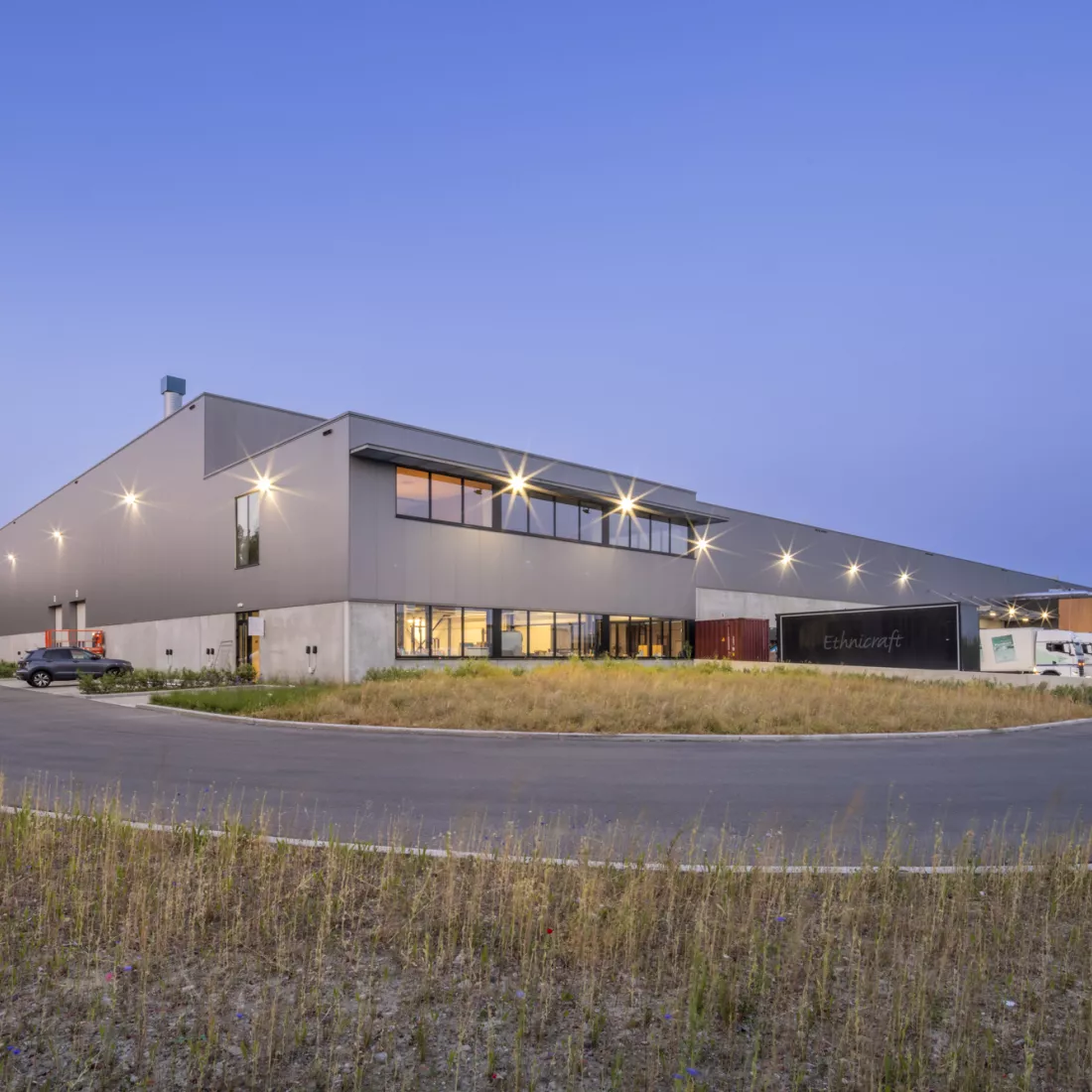 Zijaanzicht Ethnicraft. Het gebouw bestaat uit beton en staal. Aan de onderkant heeft het een betonlook en aan de bovenkant zijn grijze platen. Rechts bevindt zich een dock. Deze afbeelding is gemaakt toen het donker werd buiten.