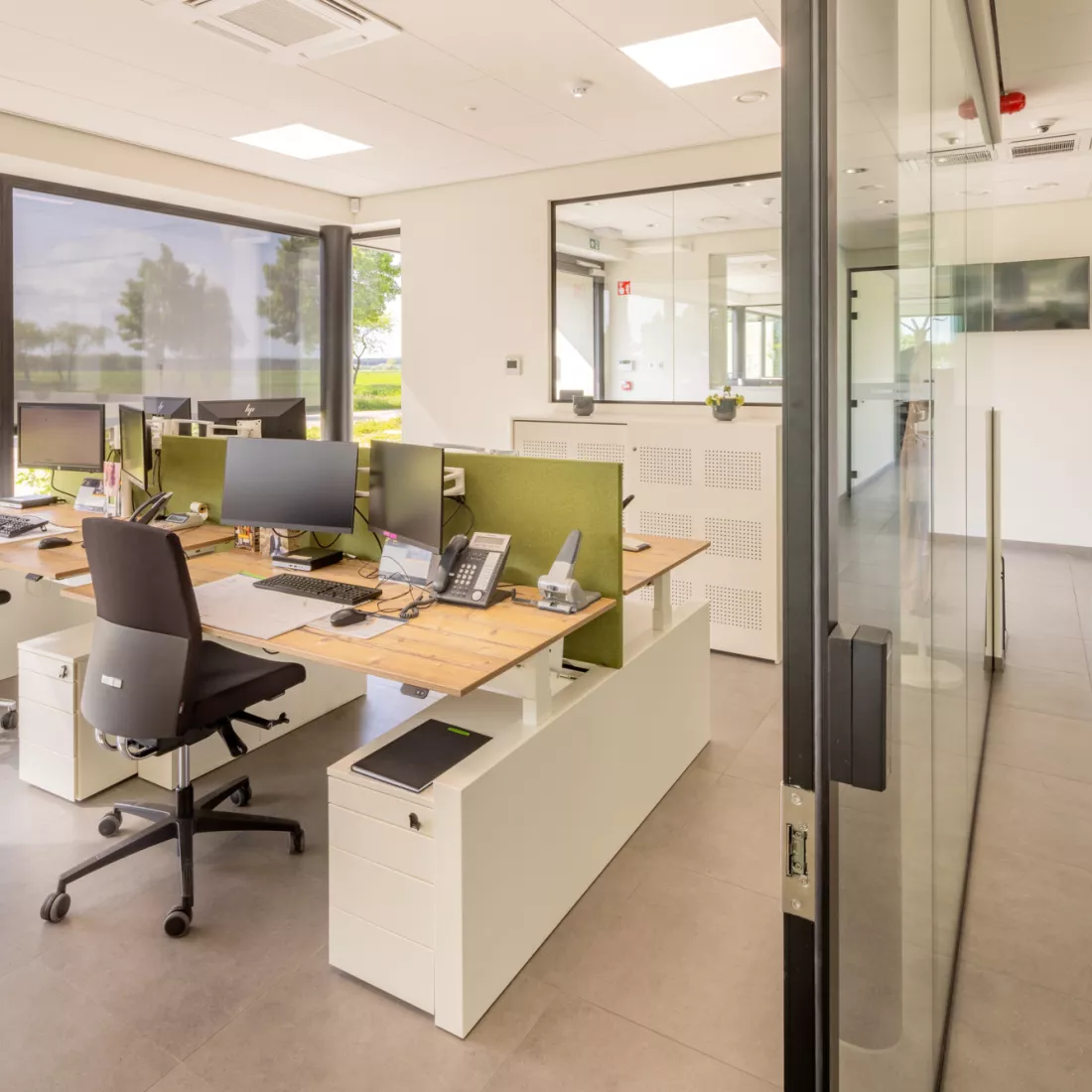 Binnenkant kantoor Omnicol. Links is er een bureel met vier bureaus. Tussen de bureaus staan groene wanden. Rechts is er een gang.