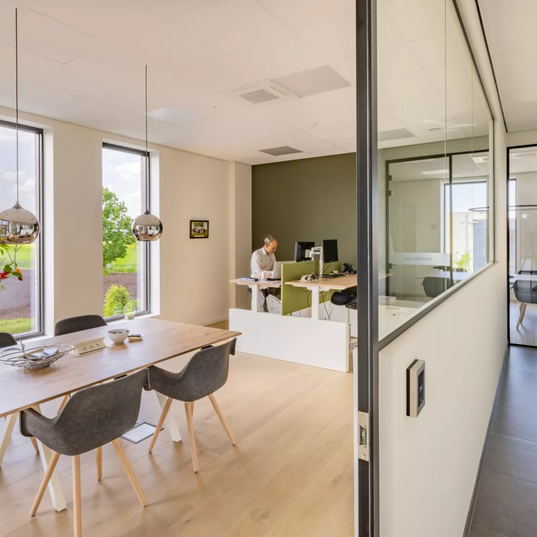 Binnenkant bureel Omnicol. Rechts in het kantoor staan twee bureaus tegenover elkaar met een groene wand ertussen. De bureaus staan tegen een groene muur. Links in het kantoor staat een rechthoekige tafel met vier tafels er rond.