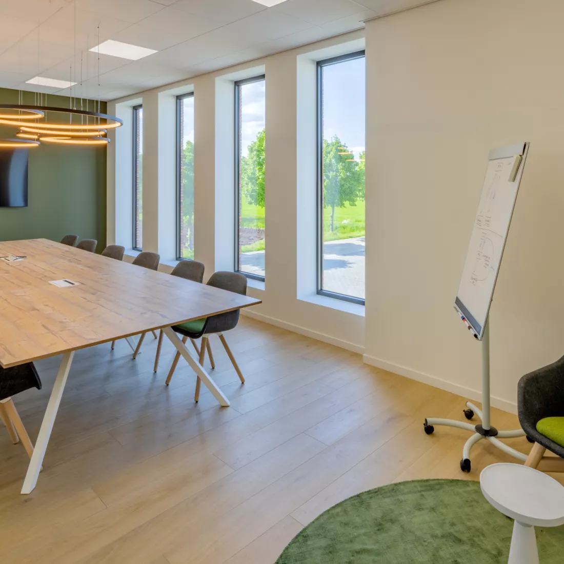 Binnenkant vergaderruimte Omnicol. Er staat een lange rechthoekige tafel met veel stoelen rond. De muur links heeft een groene accentkleur. Hier hangt ook een TV tegen. Er zijn vier lange, smalle ramen. Rechts staat een mobiel whiteboard.