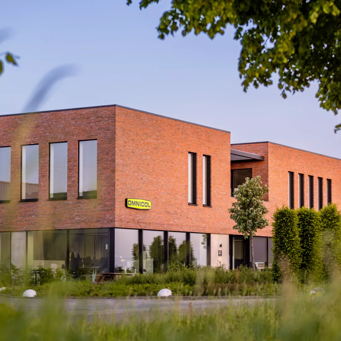 Zijaanzicht kantoor Omnicol. Voor het kantoor staat veel groen zoals een boom. Bij de opbouw van het kantoor is staal gebruikt. Het kantoor heeft twee verdiepingen. De buitenkant heeft een rode steen. Onderaan zijn heel veel grote ramen. Bovenaan zijn smallere ramen. De ramen hebben een zwarte omranding. Het logo van Omnicol hangt ook tegen het kantoor.