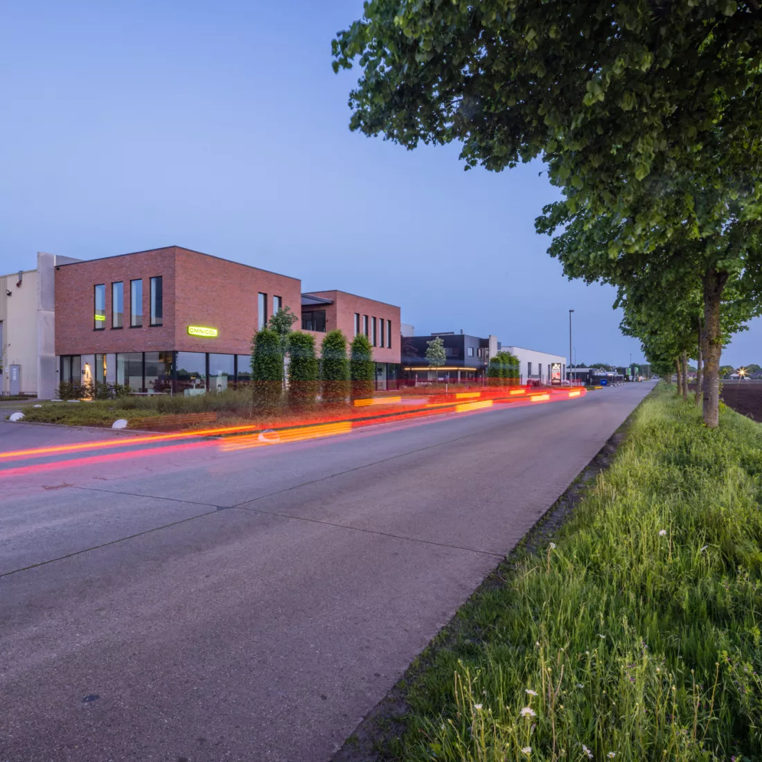 Zijaanzicht kantoor Omnicol. Voor het kantoor staat veel groen zoals een boom. Bij de opbouw van het kantoor is staal gebruikt. Het kantoor heeft twee verdiepingen. De buitenkant heeft een rode steen. Onderaan zijn heel veel grote ramen. Bovenaan zijn smallere ramen. De ramen hebben een zwarte omranding. Het logo van Omnicol hangt ook tegen het kantoor.