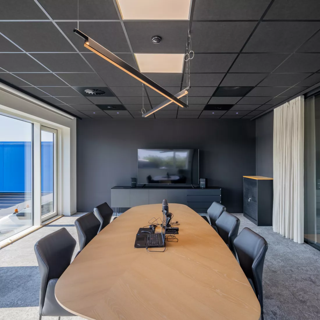Binnenkant vergaderruimte in het kantoor van Scanfor. De muren en het plafond zijn zwart. Links bevindt zich een groot raam. Tegen de muur hangt een TV met TV meubel. In het midden staat een houtentafel met zes zwarte stoelen.
