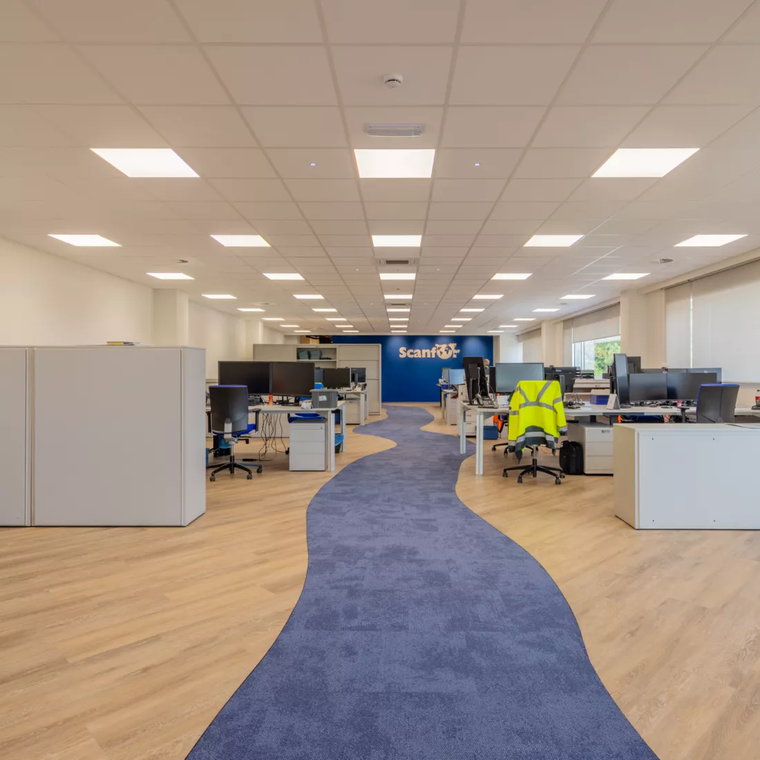 Binnenkant kantoorruimte Scanfor. In het midden ligt een kronkel weg dat bestaat uit blauw tapijt. Links en rechts hiervan staan grijze bureaus en kasten. De achterste muur heeft ook een blauwe kleur en bevat het logo van Scanfor. Rechts zijn er ramen.