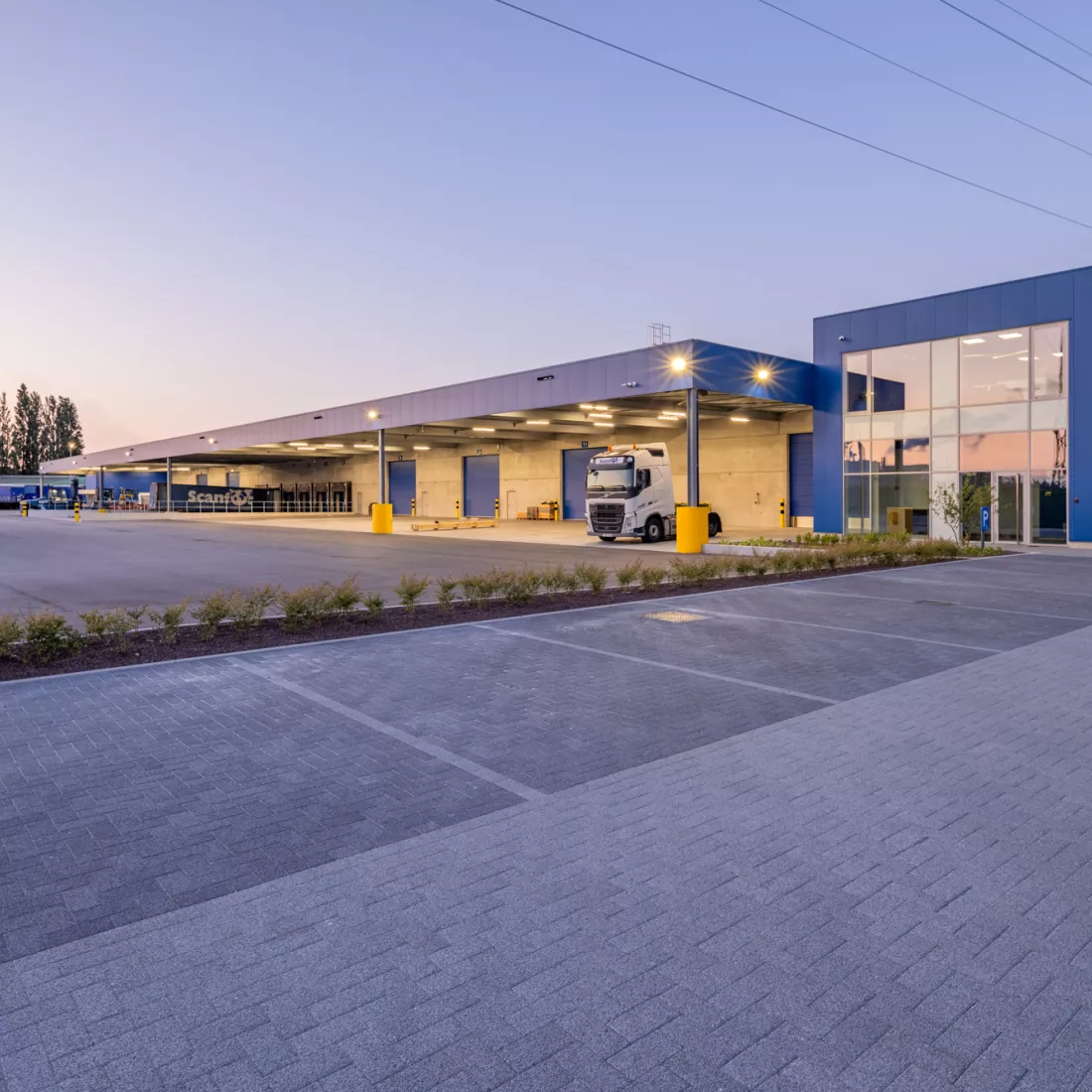 Zijaanzicht kantoor en magazijn Scanfor. Het kantoor, de poorten en het afdak aan het magazijn zijn blauw. Voor het kantoor is er parkeerplaats voorzien.
